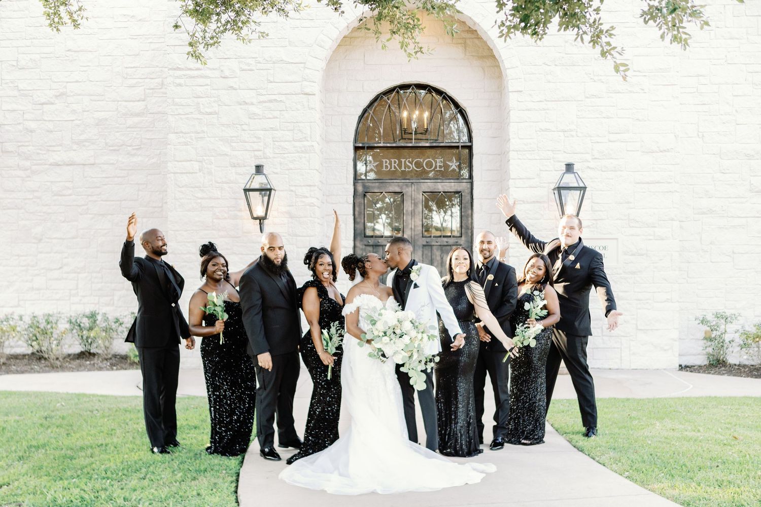 Wedding party celebrates joyfully outside a white church with arched entrance and lanterns.
