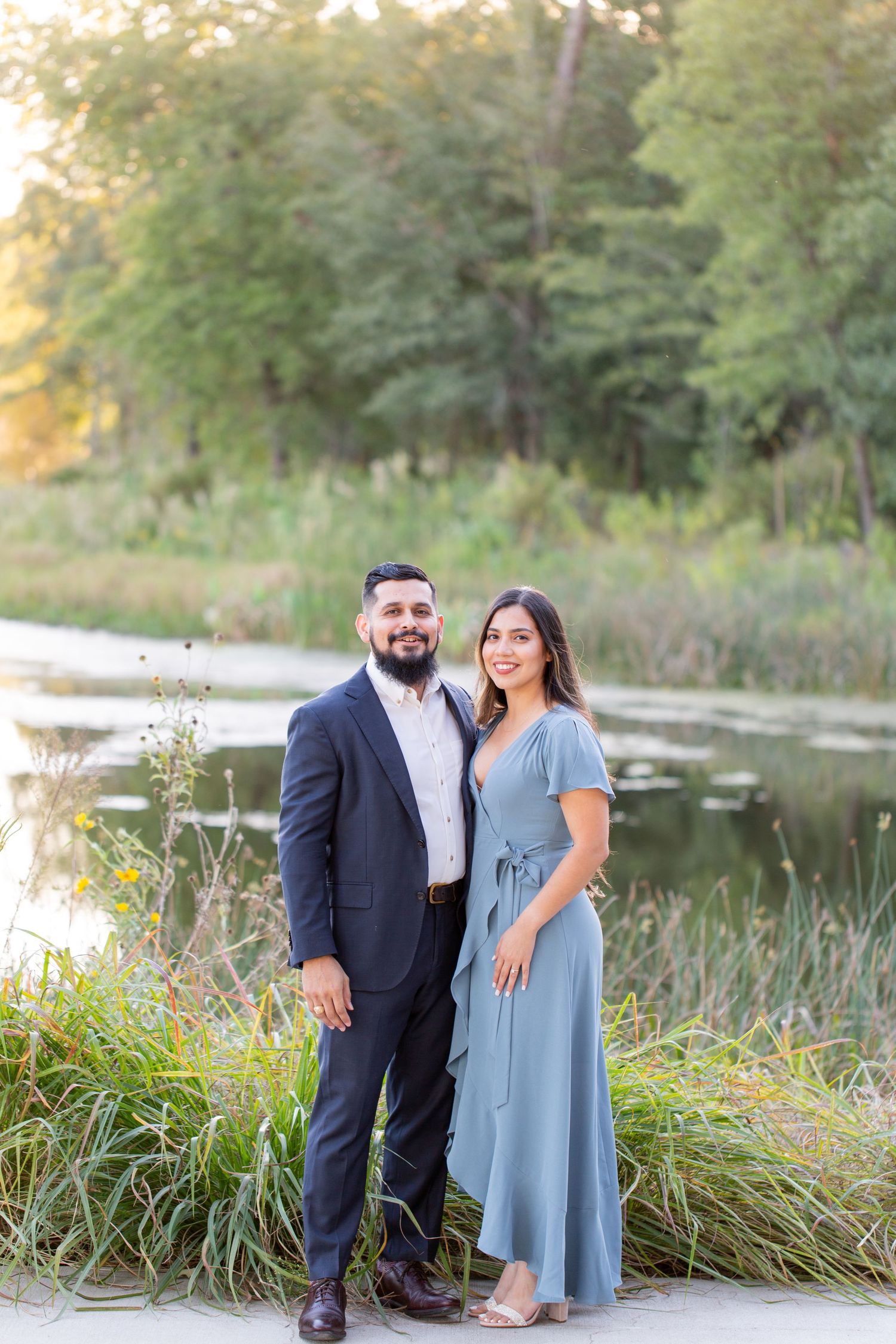 A couple poses for engagement photos by a serene lake surrounded by greenery at sunset.