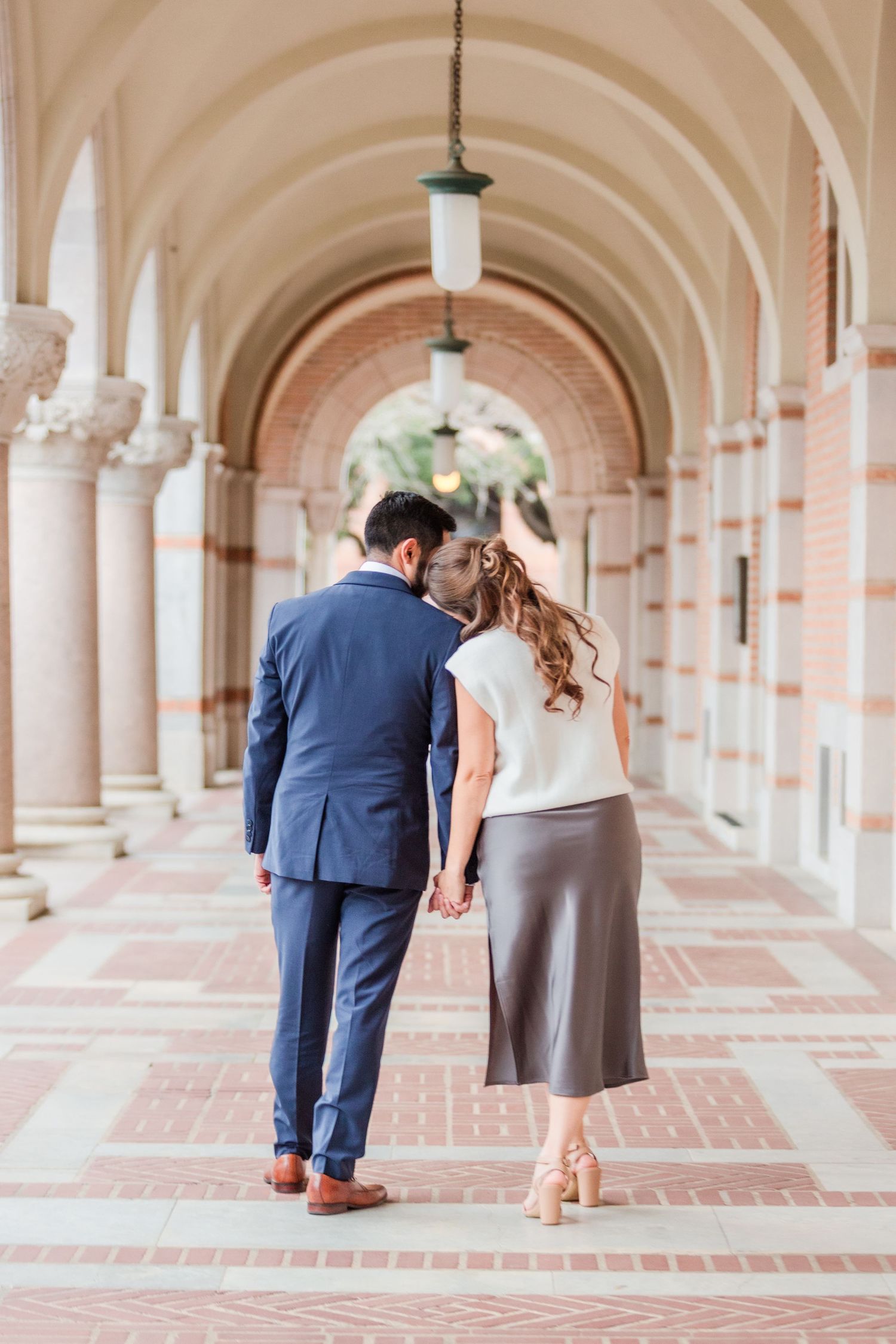 Romantic couple walks through elegant arched colonnade in formal attire.