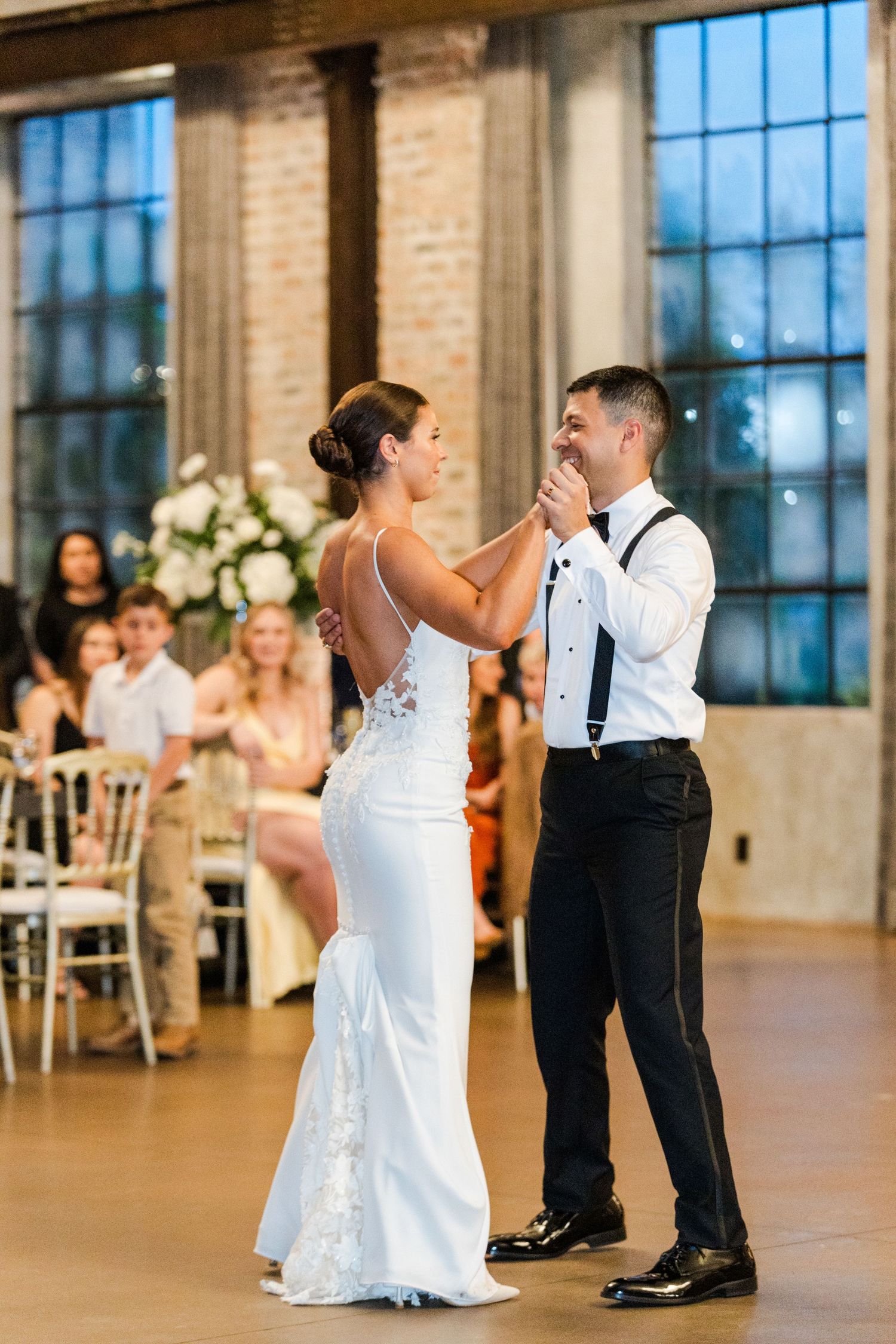 Couples enjoying their first dance in a spacious brick venue with large windows and elegant decor.