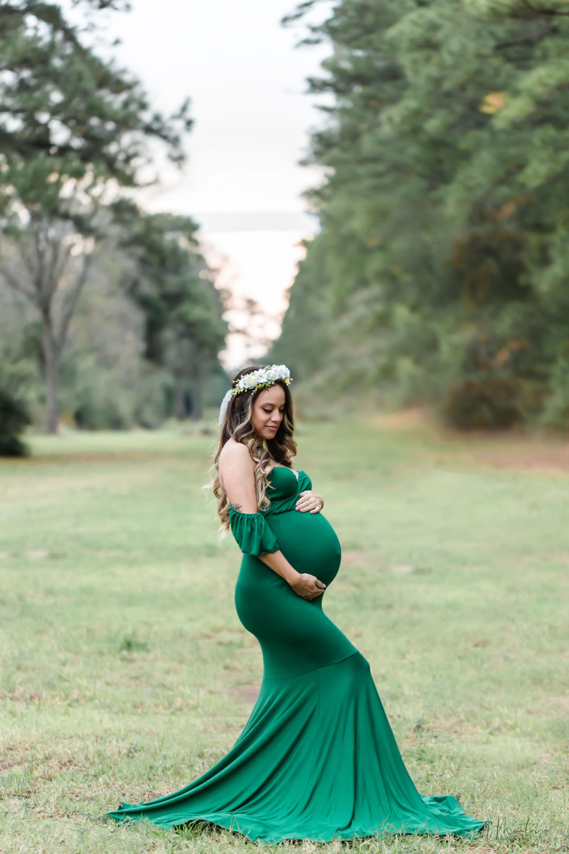 Woman in emerald green gown poses gracefully in open field during maternity shoot.