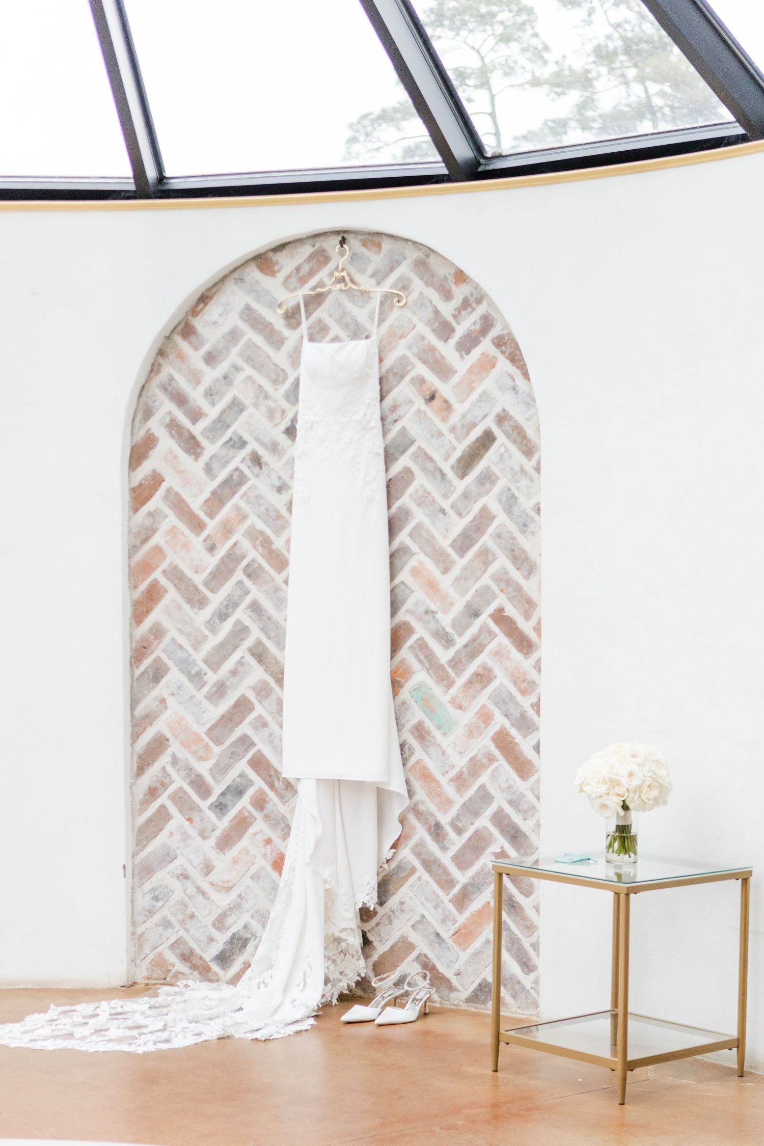 Wedding dress displayed in a bright room with herringbone brick archways and gold accent furniture.
