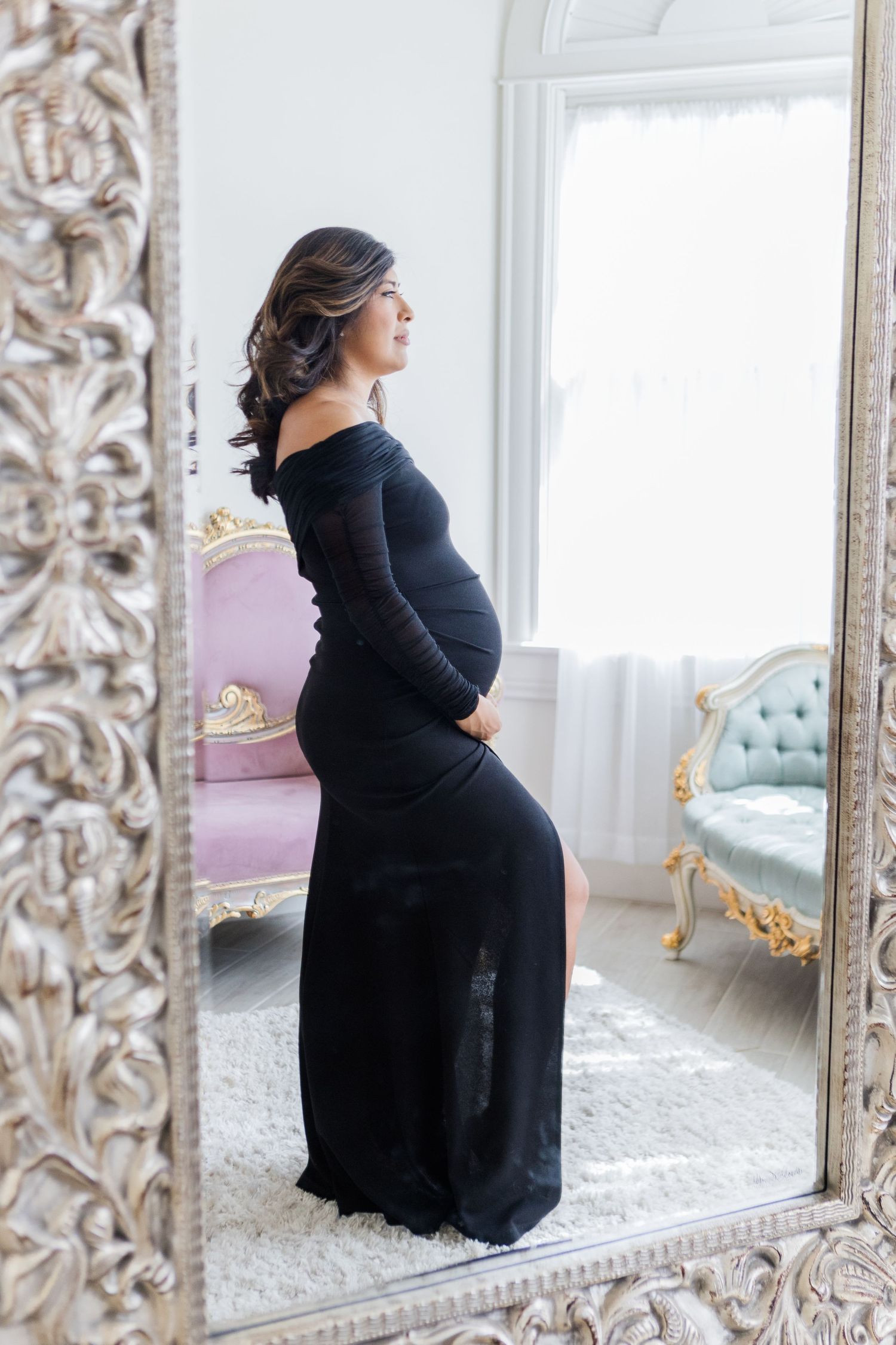 Maternity photo shoot featuring elegant black dress and ornate silver mirror in bright white room.