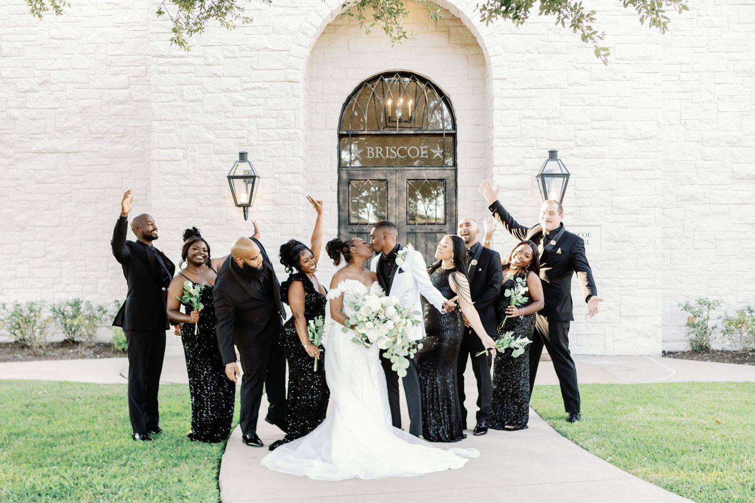 A bridal party celebrates joyfully together on the grounds of an elegant venue with white stone walls and greenery.