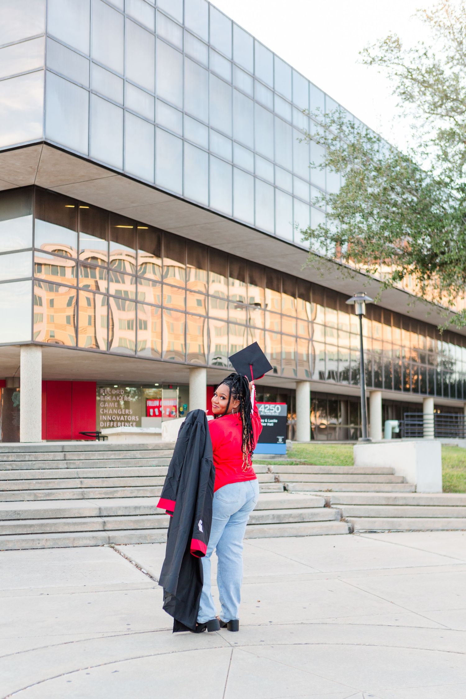 A young woman in a red shirt holds graduation cap and gown outside a modern university building.