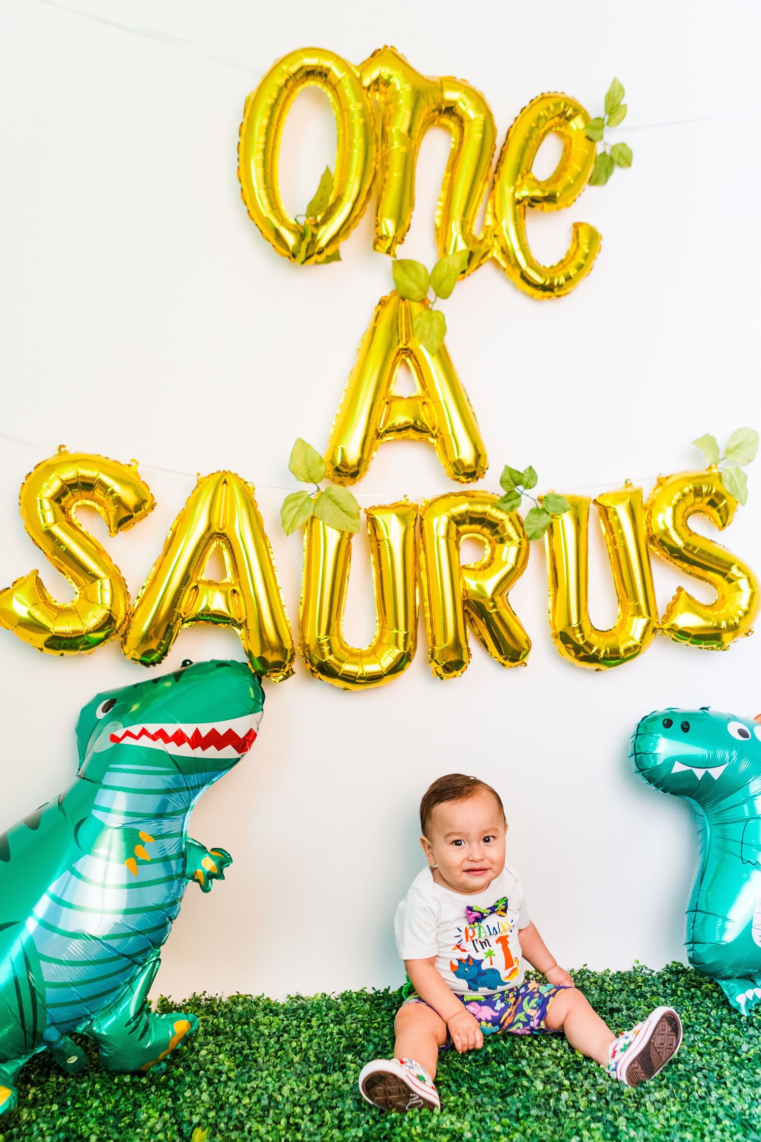 Dinosaur themed birthday party setup with teal balloon decorations and gold lettering against a white wall.