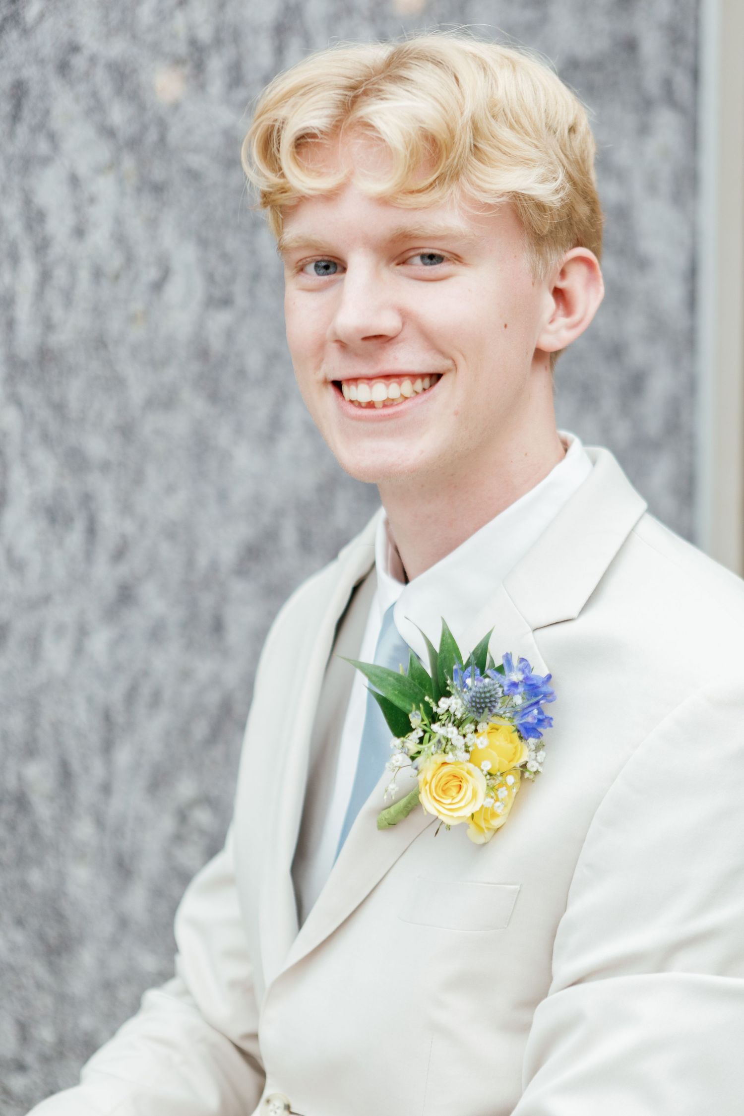 Person in white suit with yellow and blue floral boutonniere smiles against grey background.