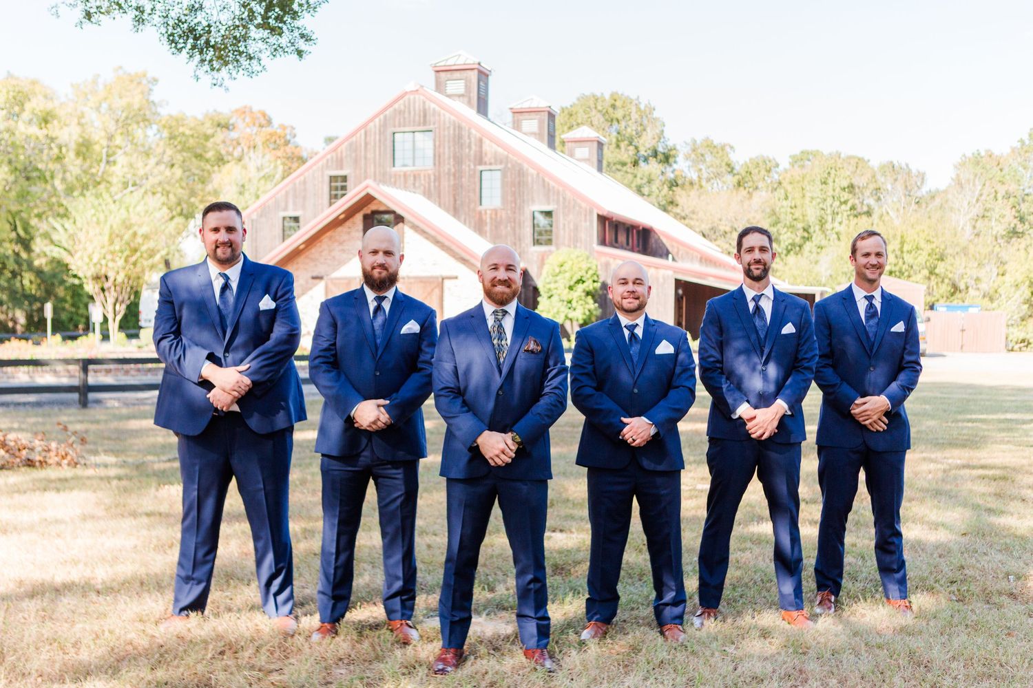 Wedding party members in navy blue suits pose in front of rustic barn venue.