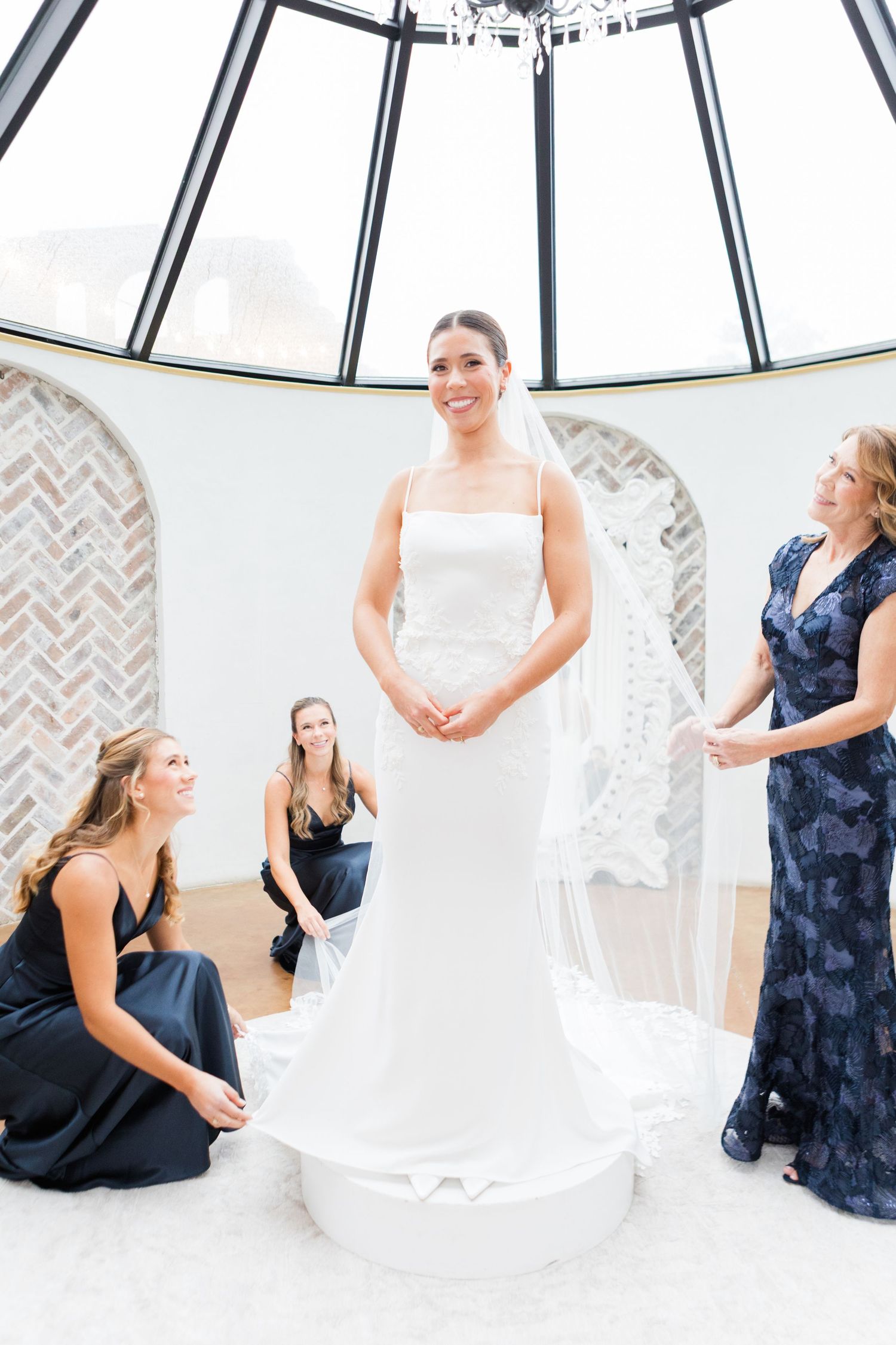 Wedding party scene with bridesmaids in navy dresses and bride in white gown in a bright indoor setting.