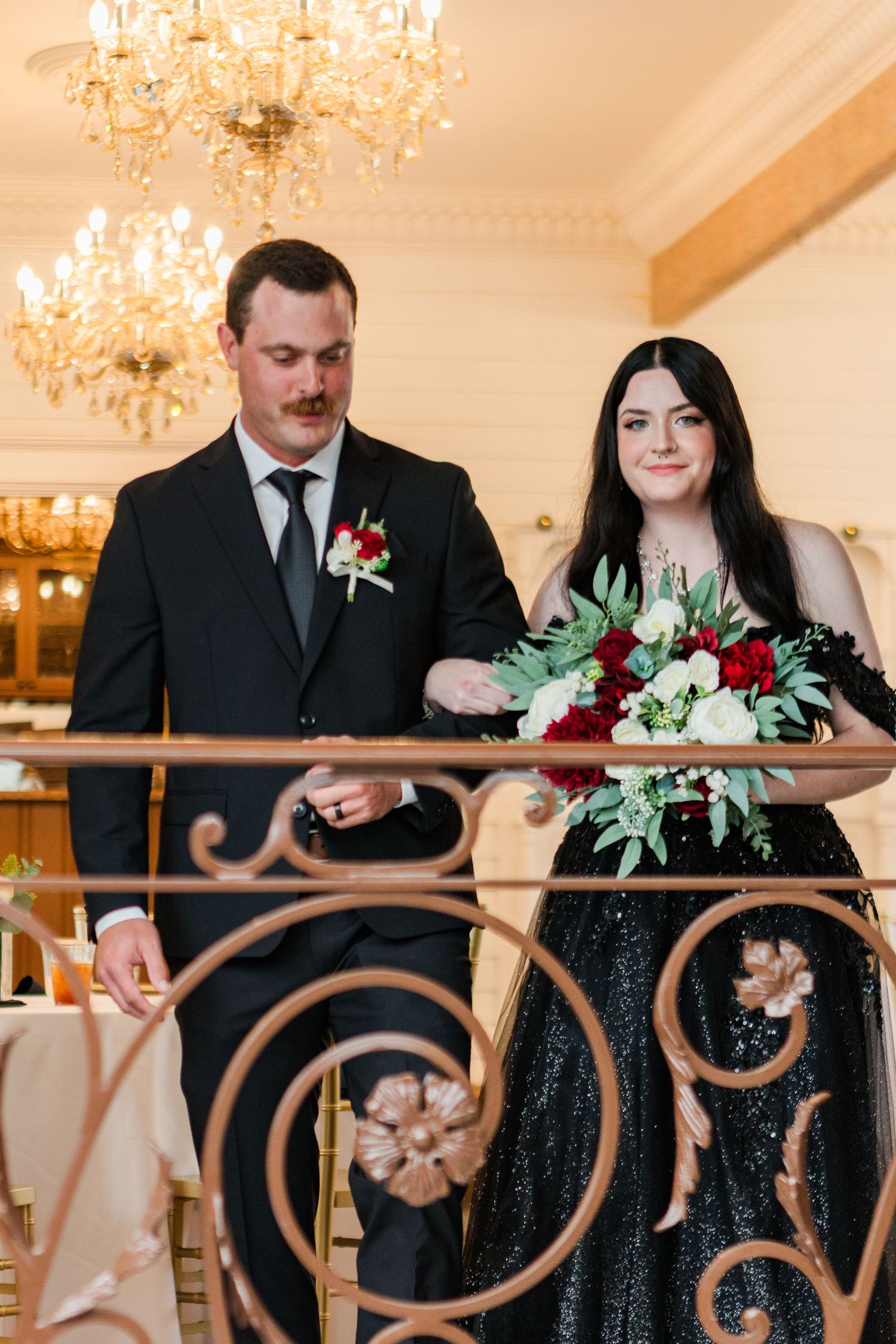 Wedding couple in formal black attire posing near ornate gold railings under crystal chandeliers.