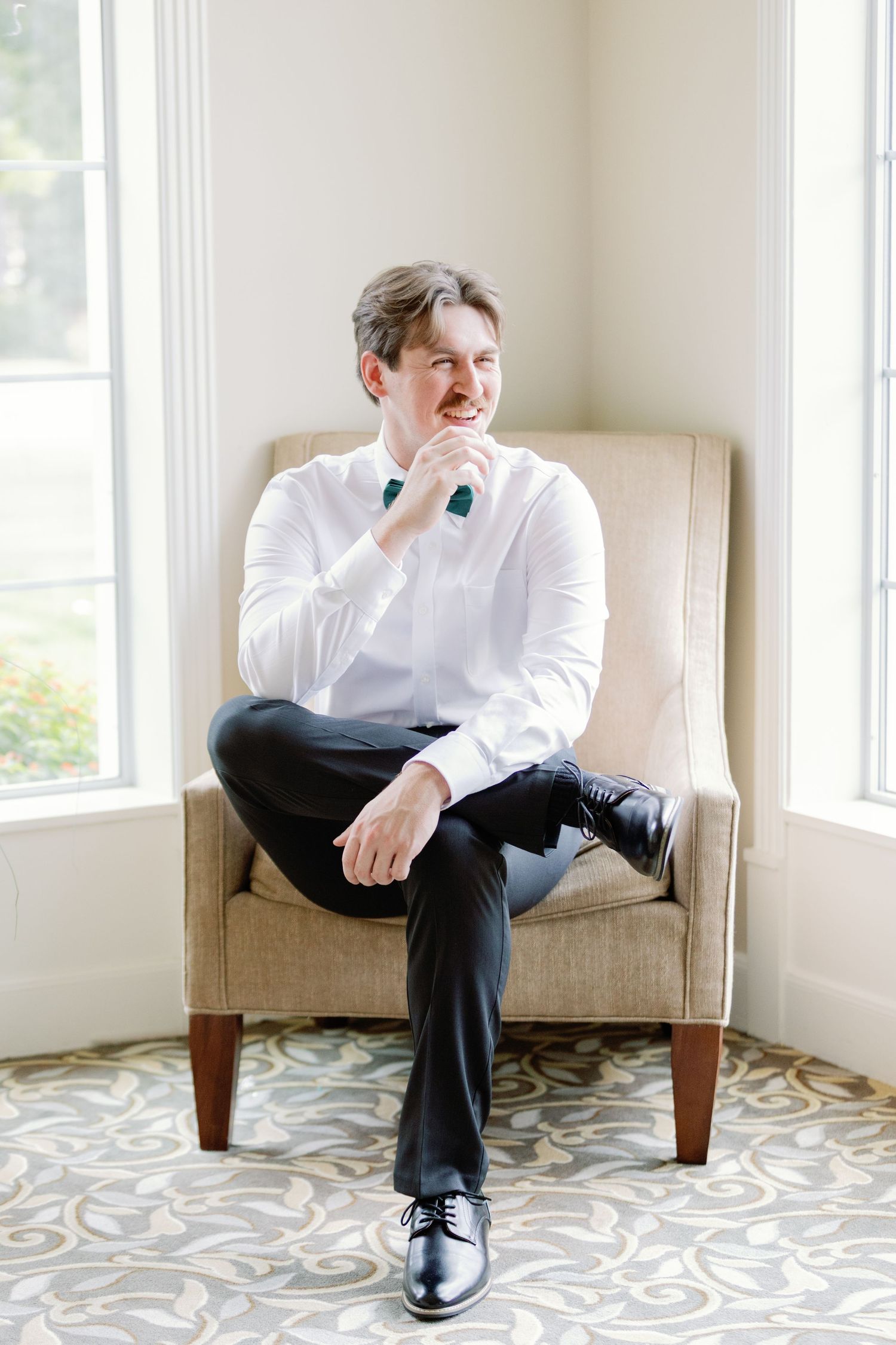 A well-dressed person sits thoughtfully in a beige armchair by a bright window.