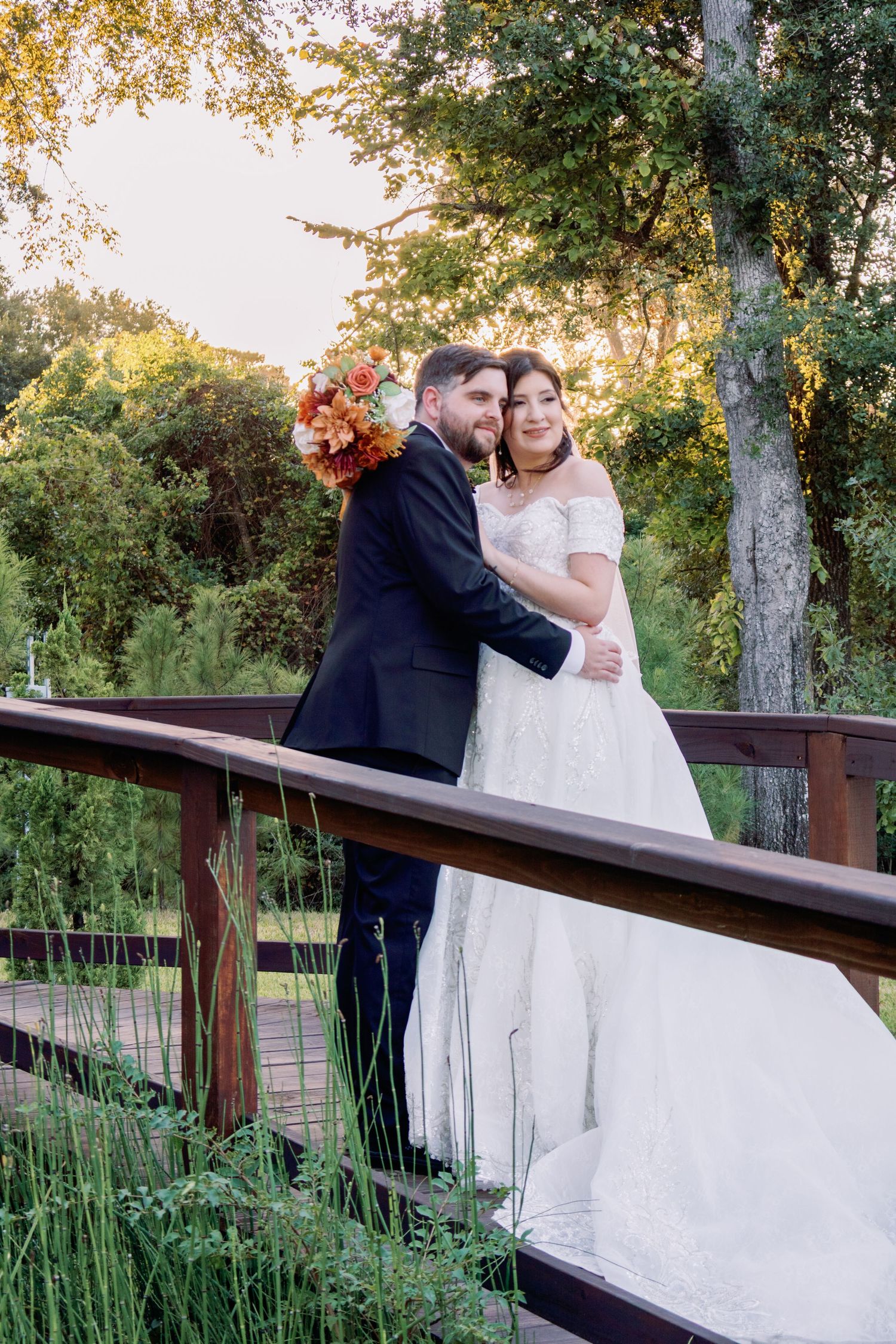 A couple embraces on a wooden bridge in a garden setting during golden hour with autumn foliage in the background.