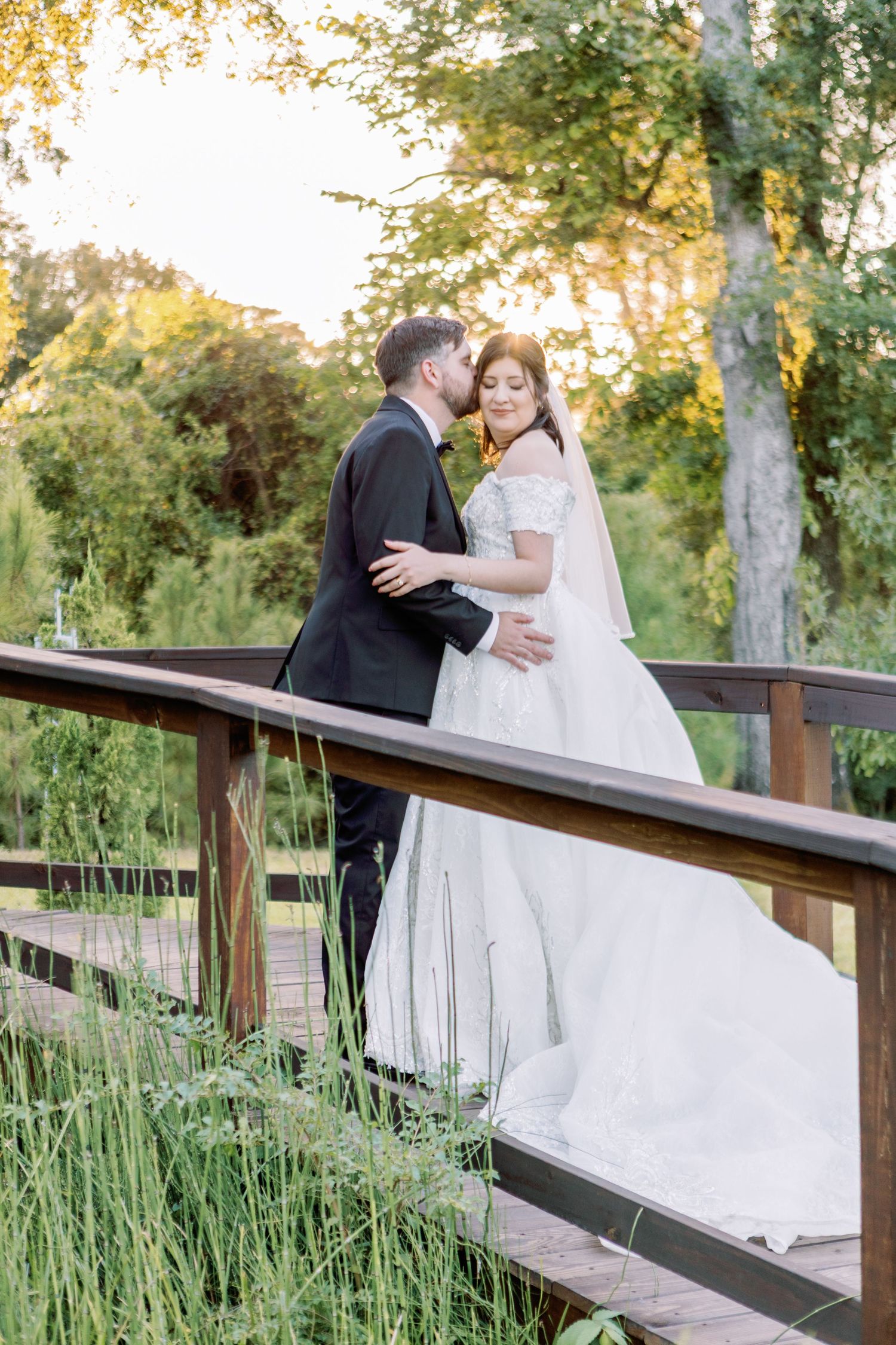 Newlyweds share intimate moment on wooden bridge surrounded by tall grass and autumn trees.