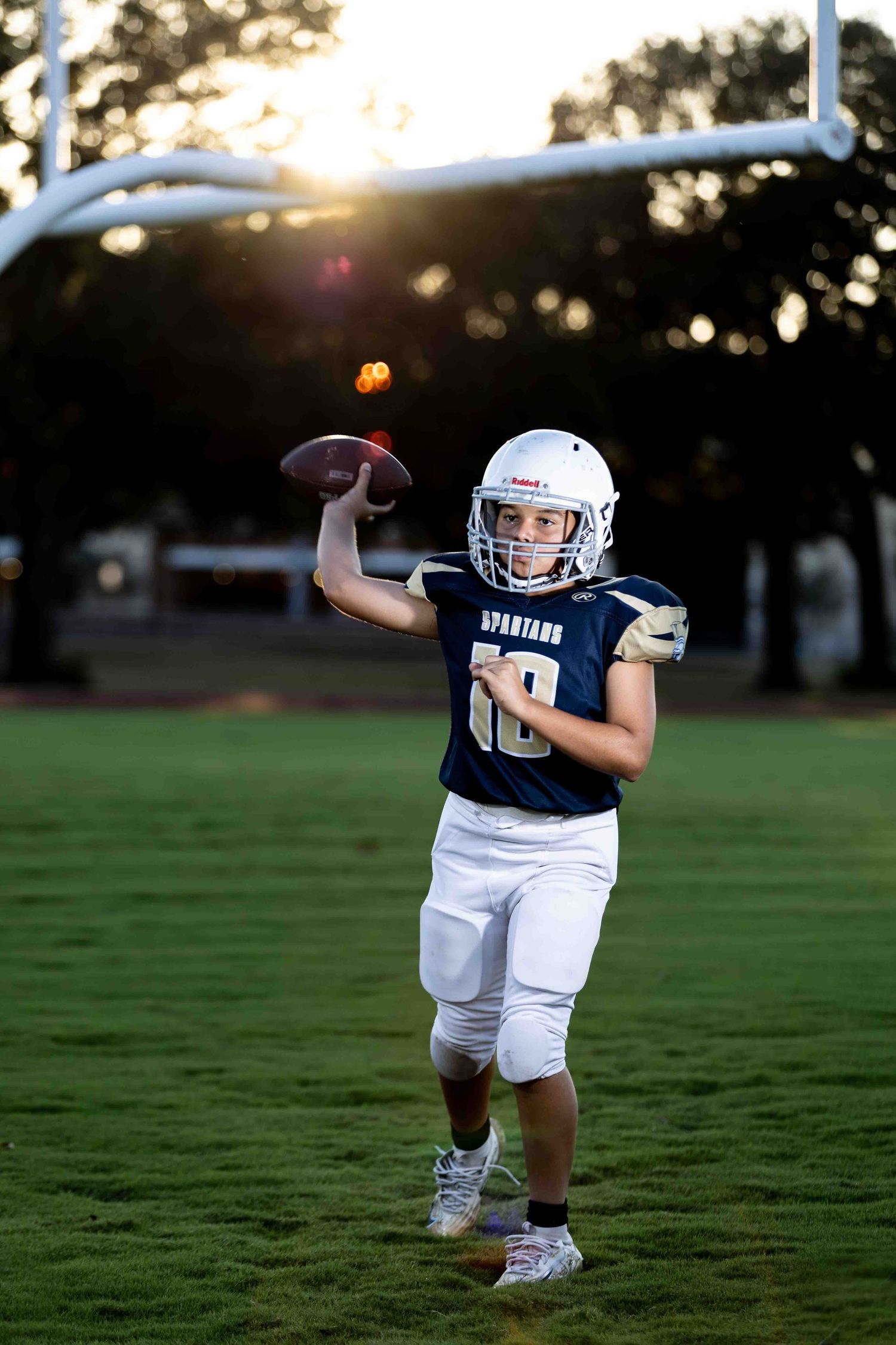 Young football player in white uniform practices throwing drills on a grass field at sunset.