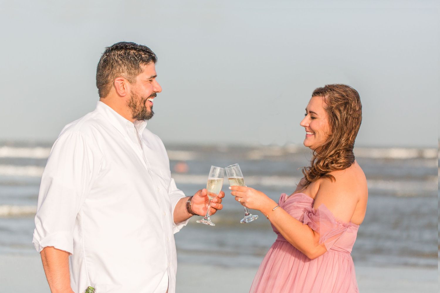 Couple toasting with drinks during beach engagement photoshoot.