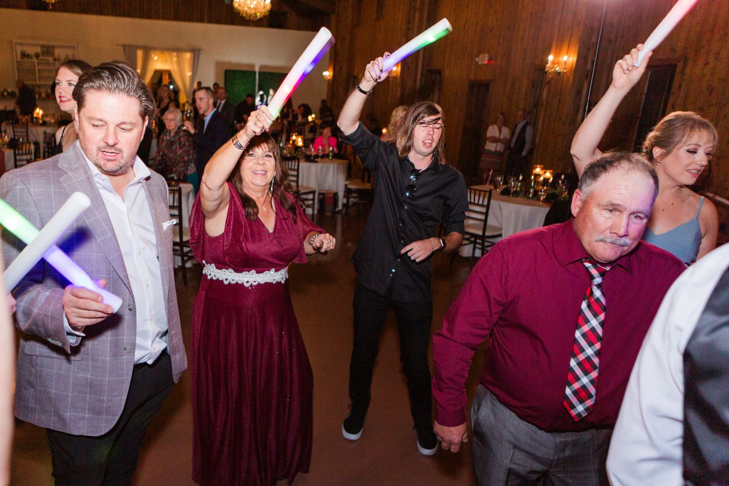 Wedding reception guests dance energetically in a rustic barn venue with mood lighting.