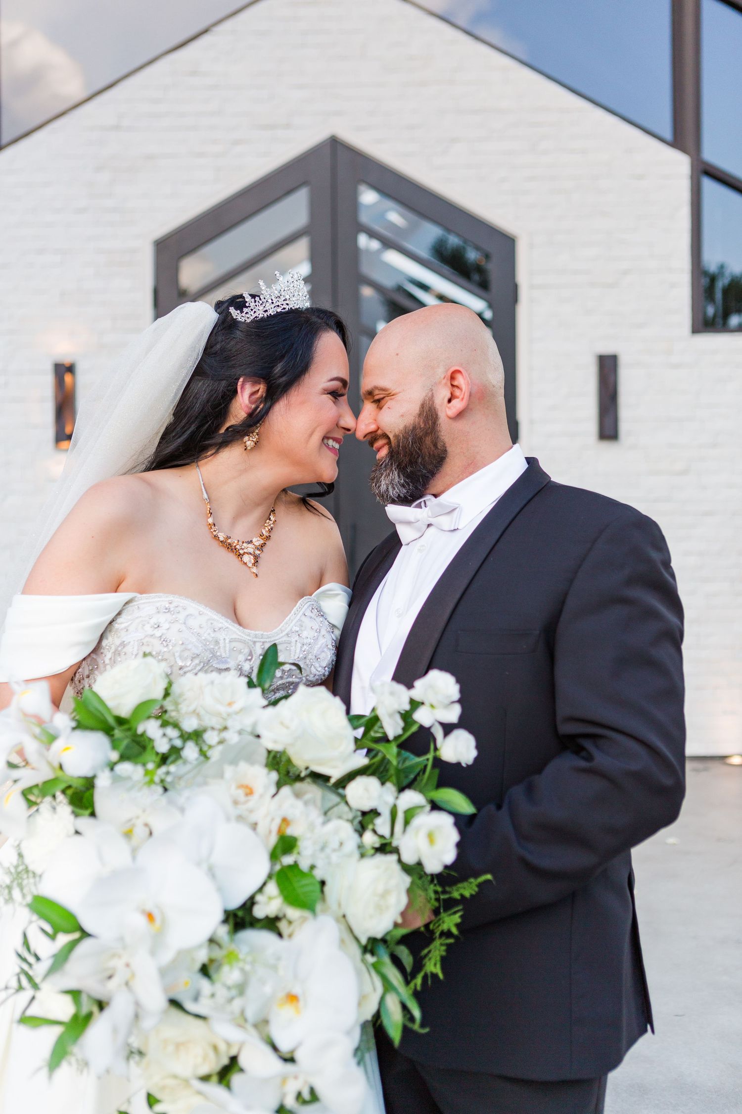 Elegant wedding couple embracing with white floral bouquet against modern white building backdrop.