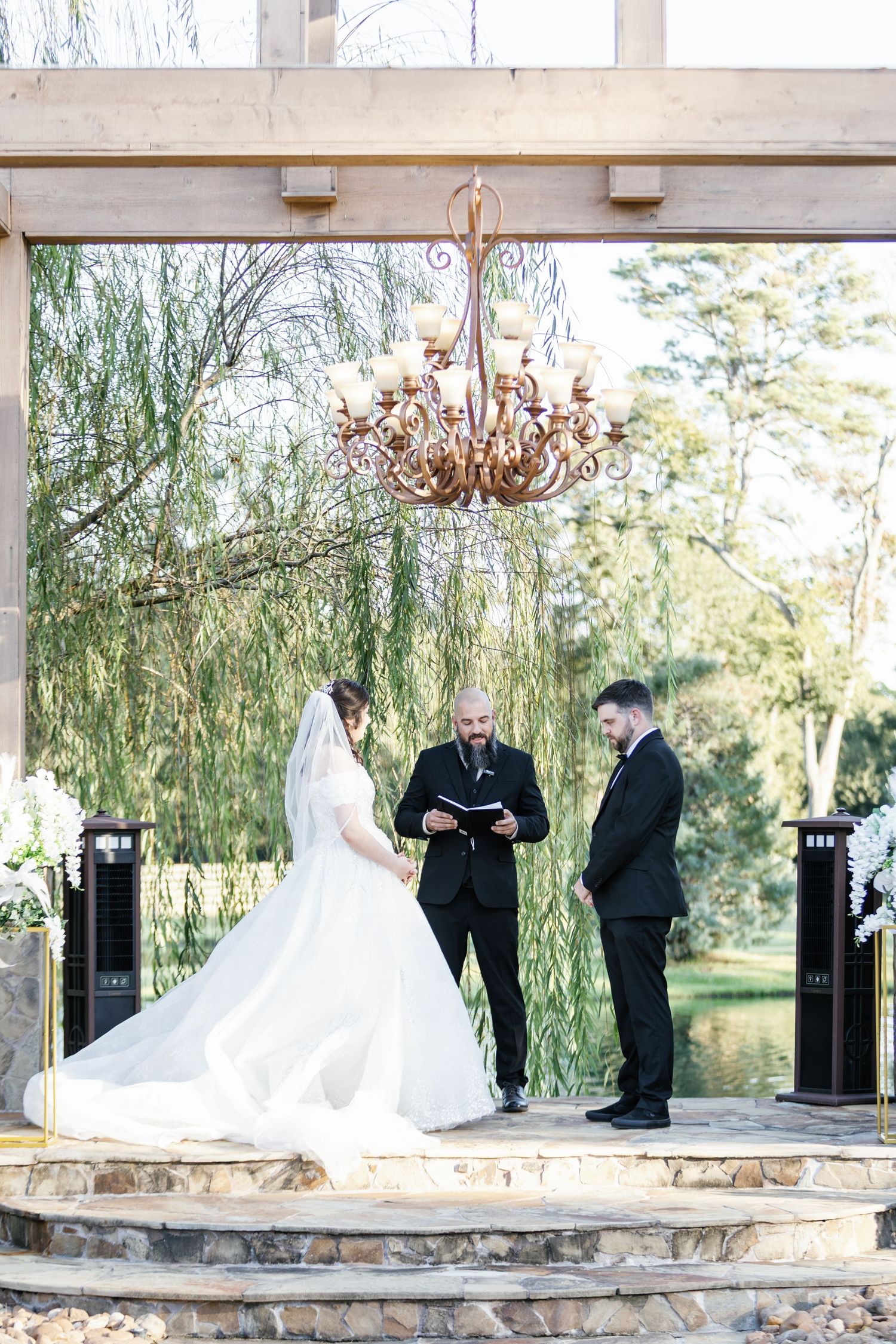 Elegant outdoor wedding ceremony setup under a wooden pergola with willow trees in the background.