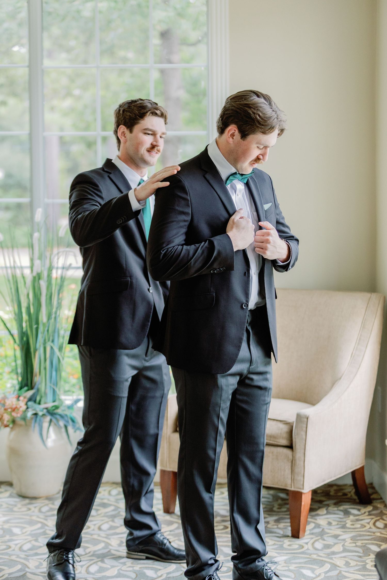 Two people in dark suits playfully pose in a bright indoor setting with patterned carpet.