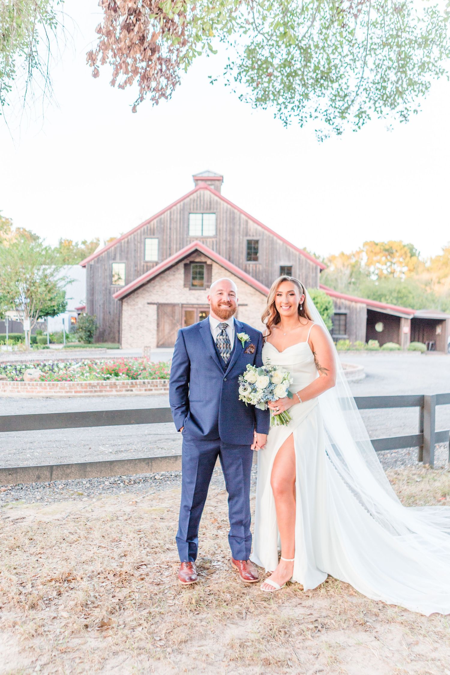 Newlyweds in blue suit and white dress posing near a rustic fence at their outdoor venue.