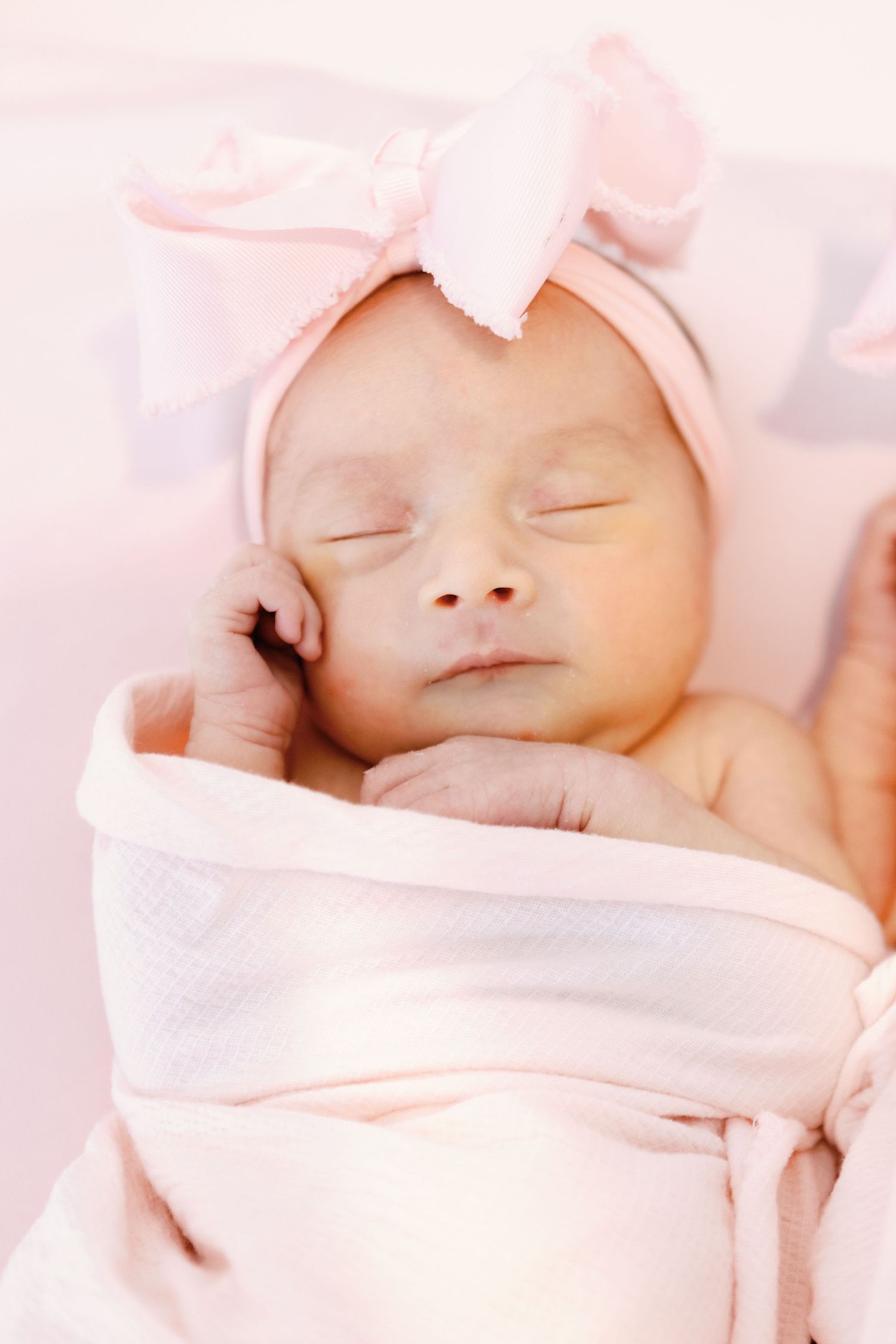 Peaceful newborn baby sleeping while wrapped in soft pink blankets and wearing a gentle bow headband.