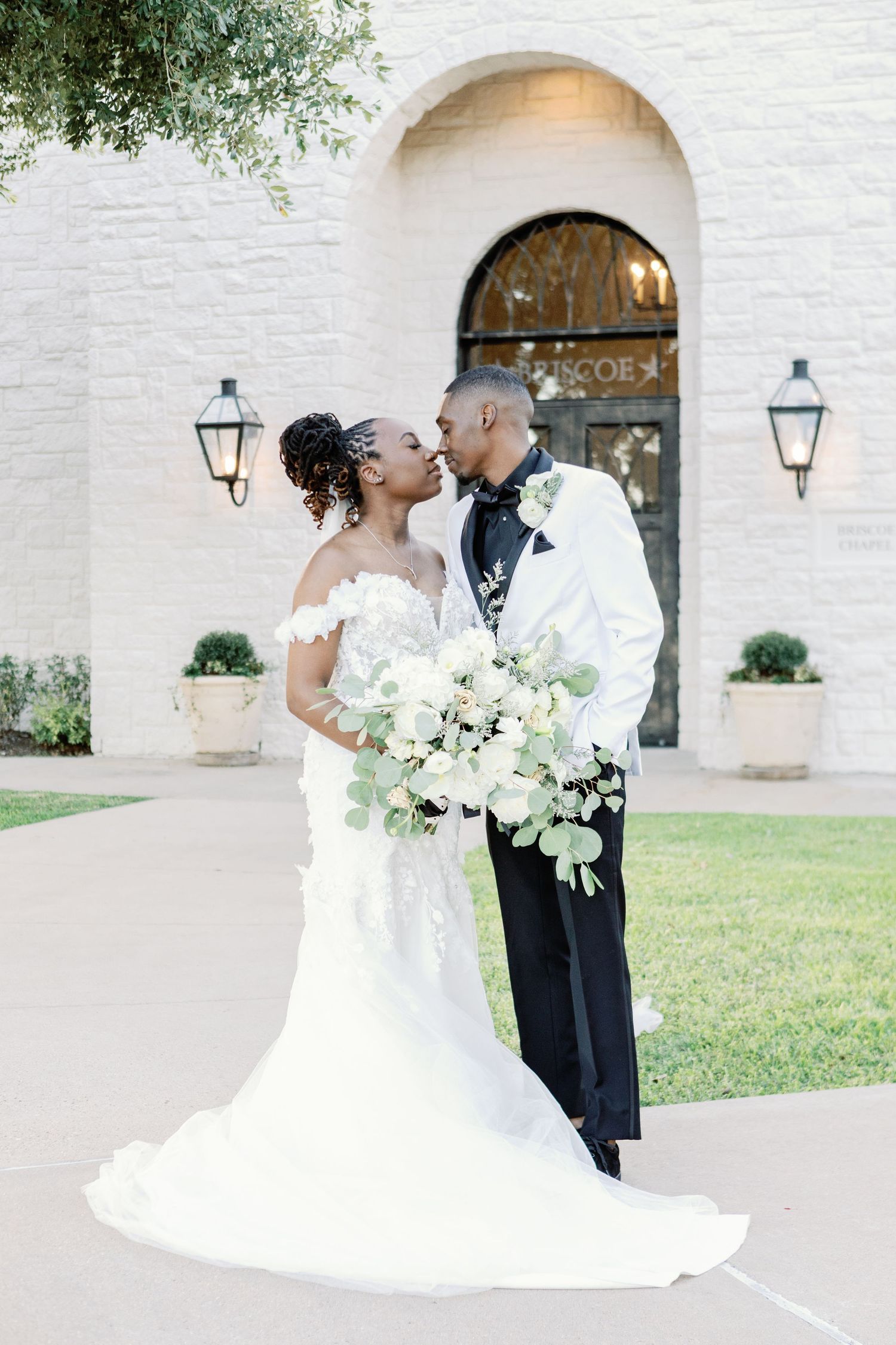 Romantic outdoor wedding portraits against white stone wall with garden lamps and natural greenery overhead.