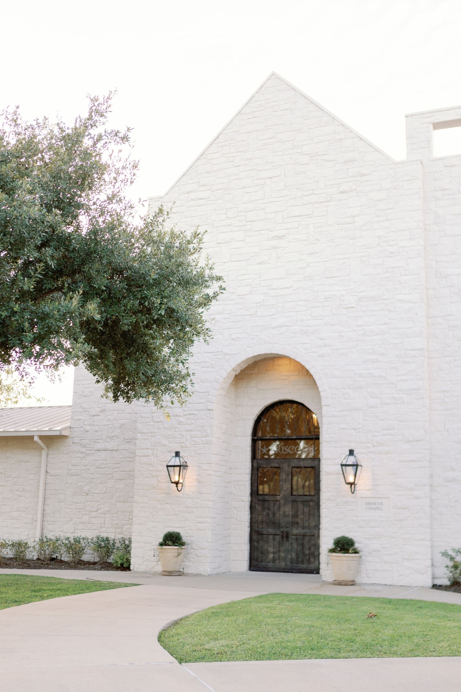Modern white stone church facade with arched doorway, wall sconces, and manicured lawn under mature oak tree.