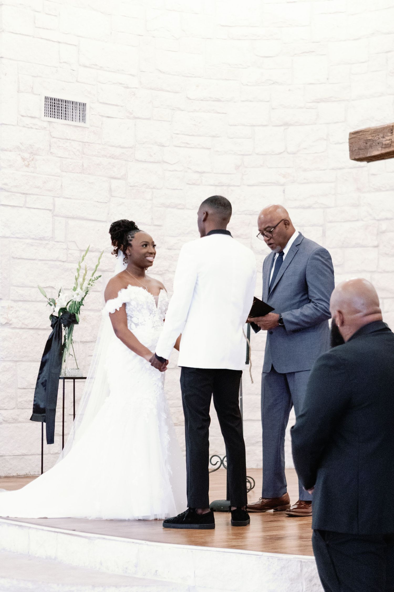 Wedding ceremony participants standing at the altar in front of a white brick wall with wooden cross.