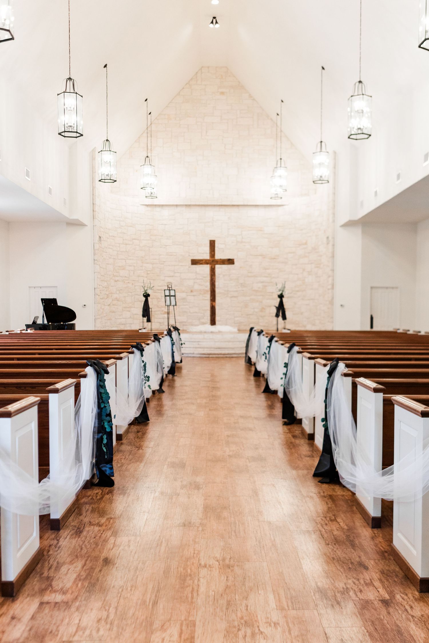Interior of a bright church chapel with wooden pews decorated with white fabric and a cross on the altar wall.