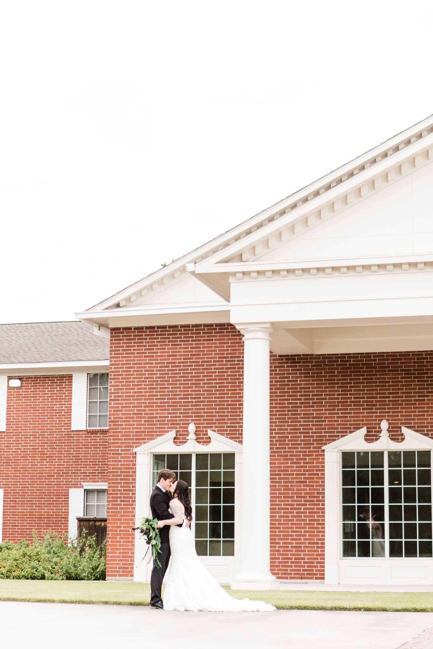 Wedding couple shares intimate moments against red brick building with white columns.