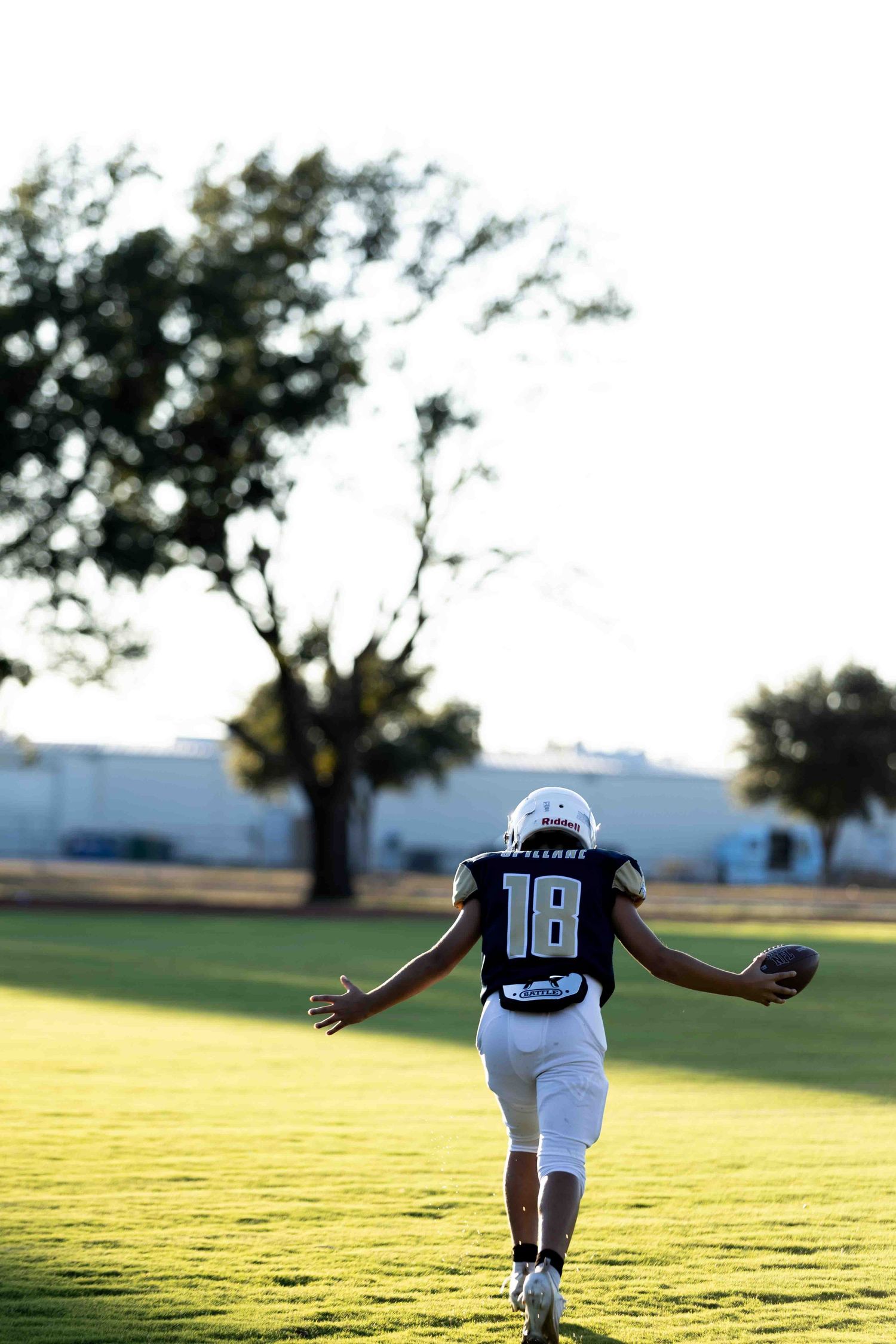 Football player in white jersey runs across sunlit grass field during golden hour practice session.