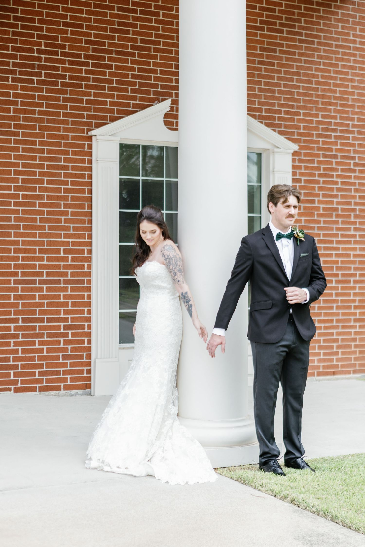 Wedding couple plays peek-a-boo around white columns against brick wall exterior.