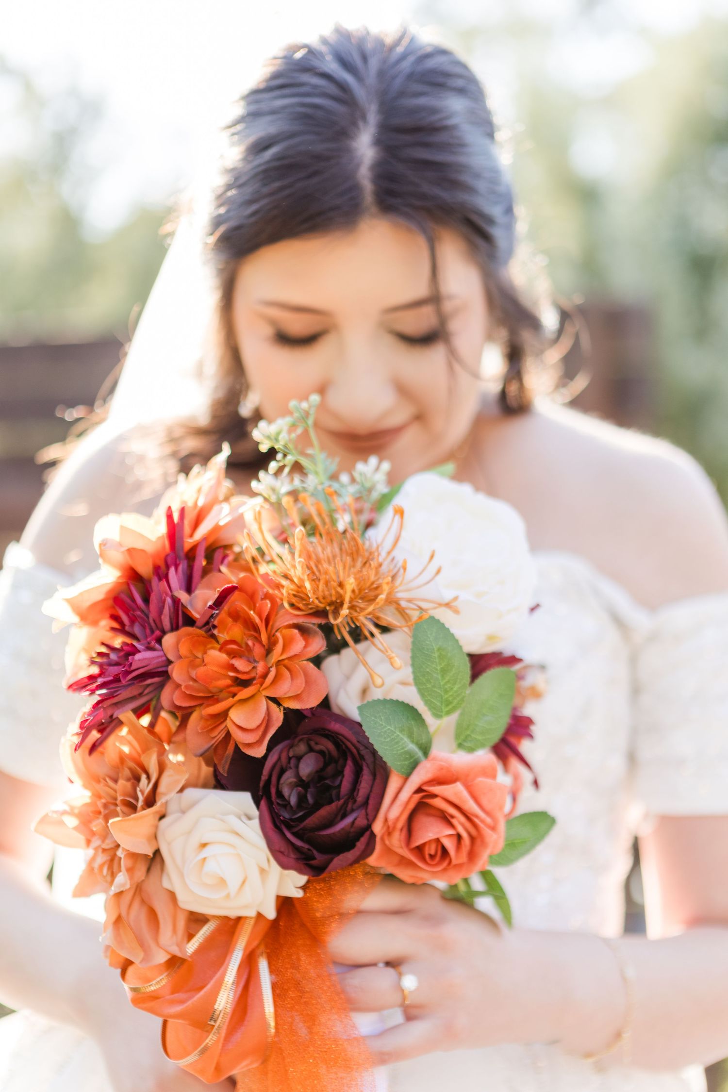 Beautiful autumn wedding bouquet with orange, burgundy and blush roses held by bride in white dress in soft sunlight.