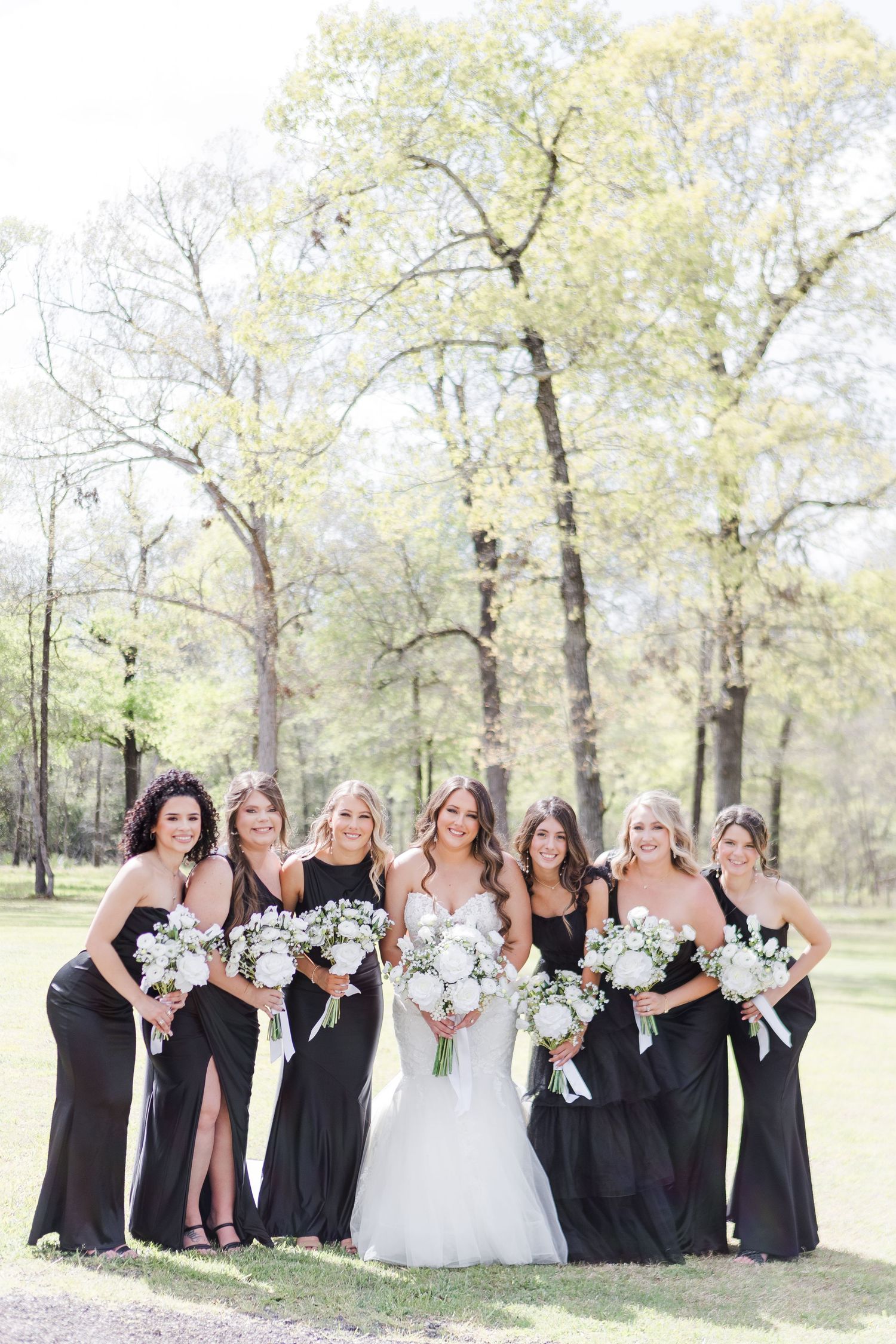 Wedding party in black dresses posing outdoors with white bouquets among spring trees.