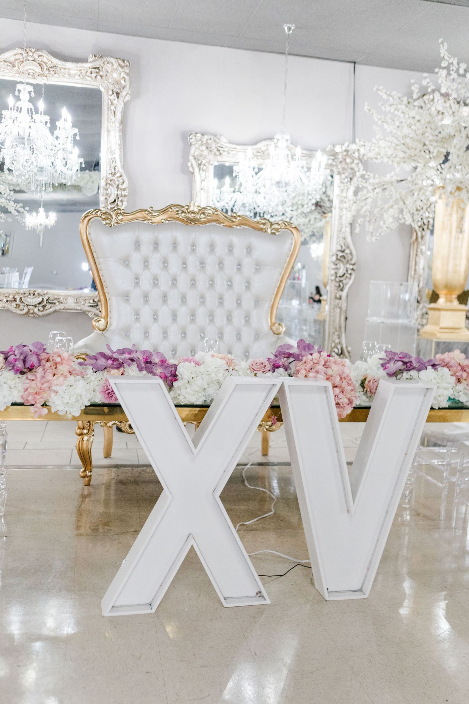 Ornate white venue with crystal chandeliers, gilded mirrors, and white Roman numeral decor.