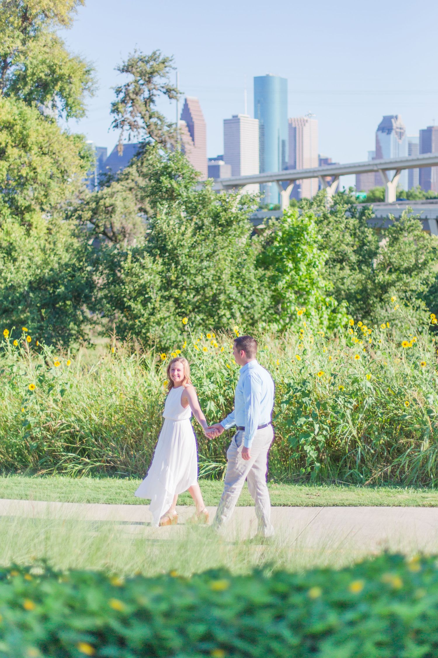 Couple strolling through lush garden with urban skyline backdrop on sunny day.