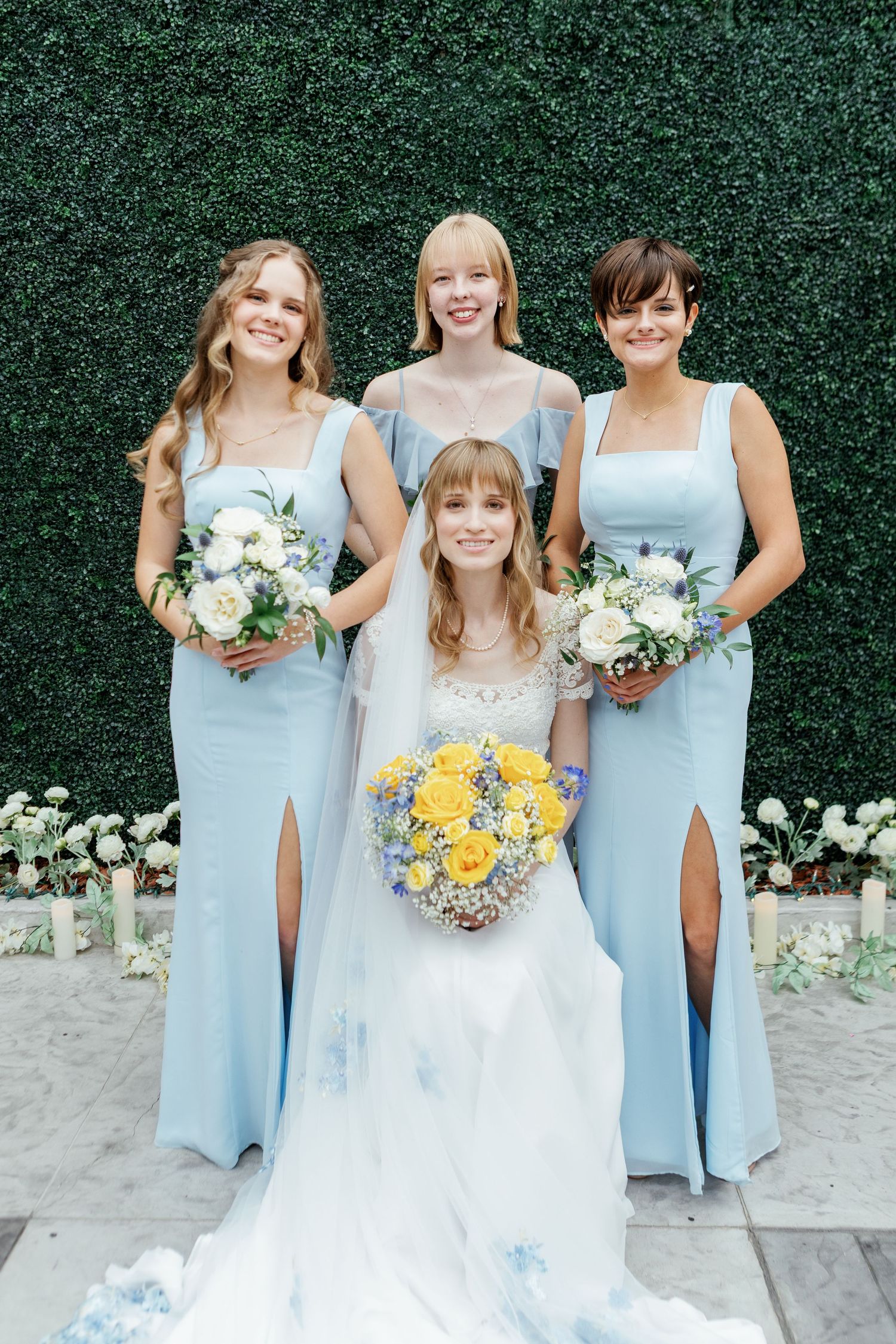 Bridesmaids in light blue dresses hold white and yellow bouquets against a green hedge backdrop at an outdoor wedding.