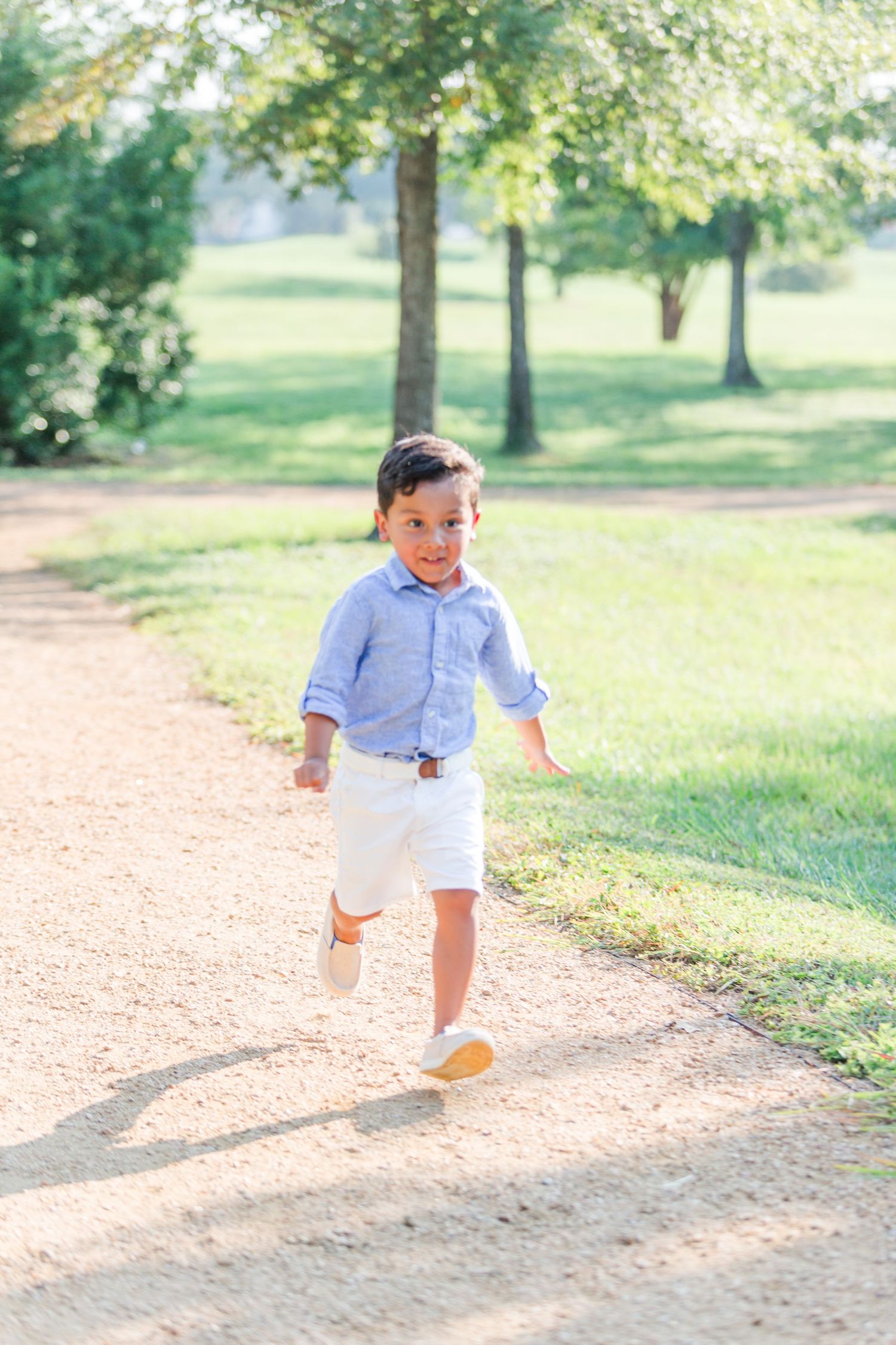 Small child in light colored outfit runs along sunny park path.