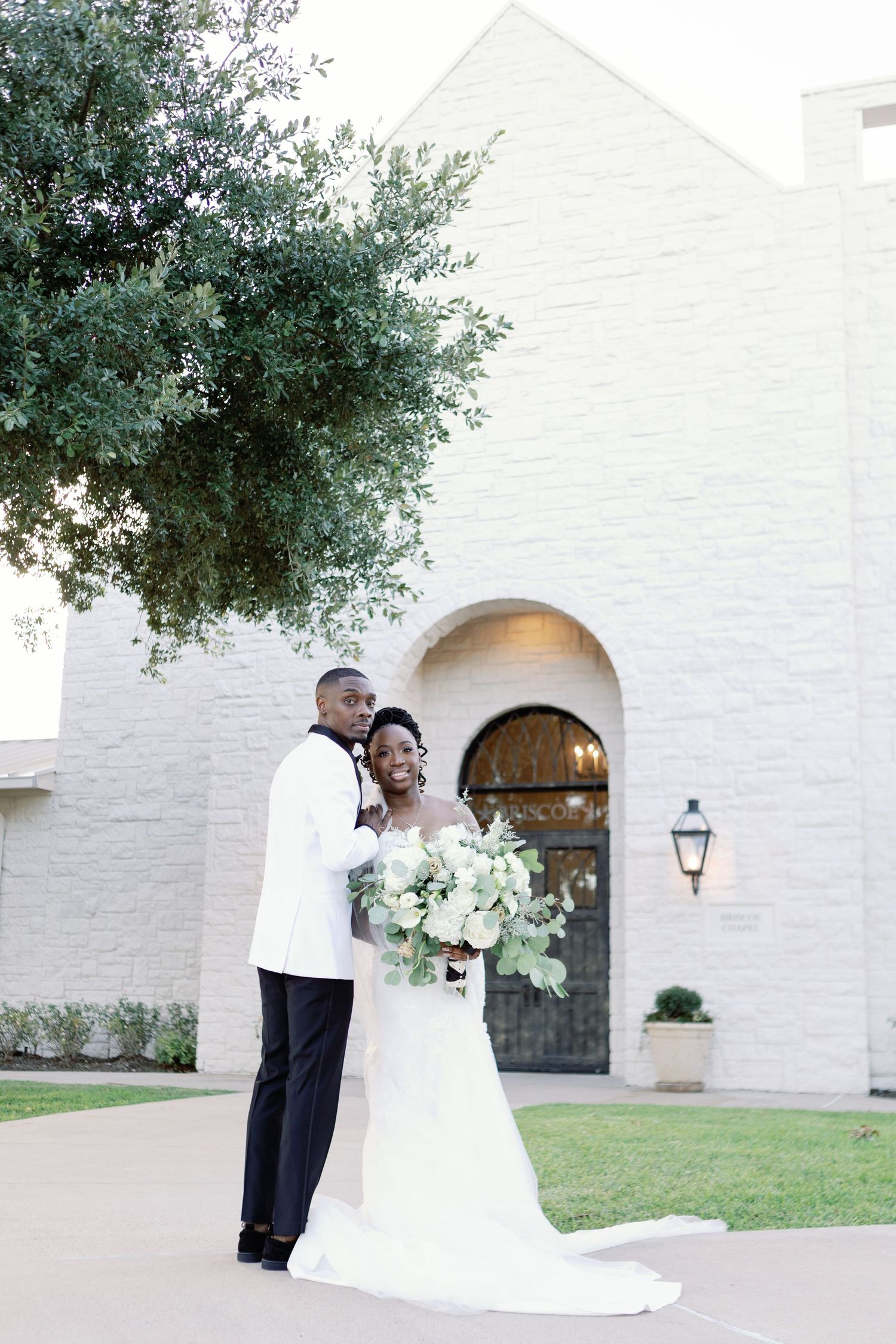 A joyful newlywed couple embracing outside a white church building with arched doorway and green foliage.