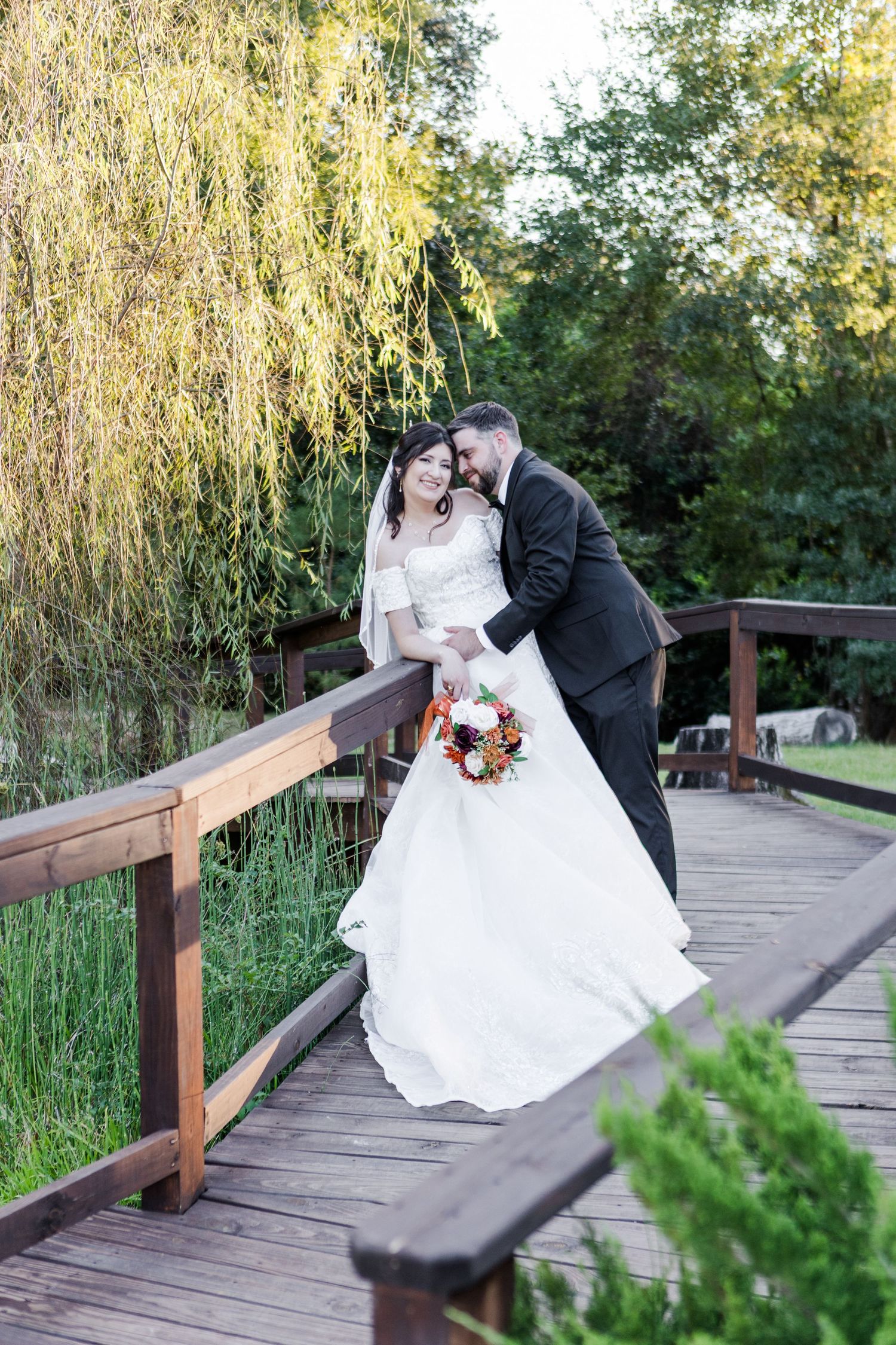 Wedding couple poses romantically on a wooden bridge surrounded by willow trees and lush greenery.