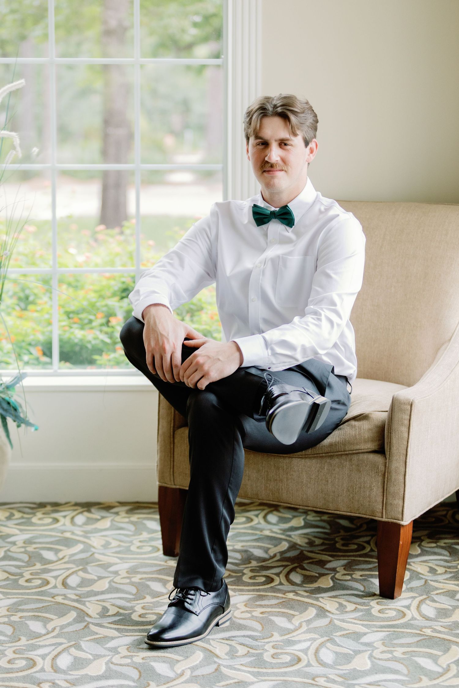 Formal attire with bow tie and dress shoes while seated in an elegant chair by the window.