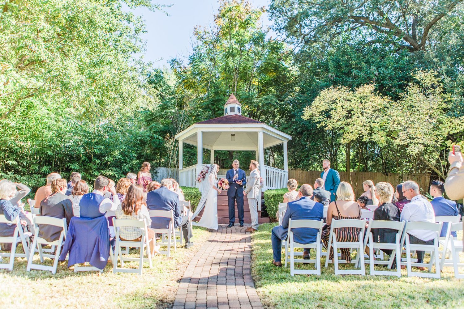 Wedding ceremony guests seated outdoors under leafy trees with a white gazebo in the background.