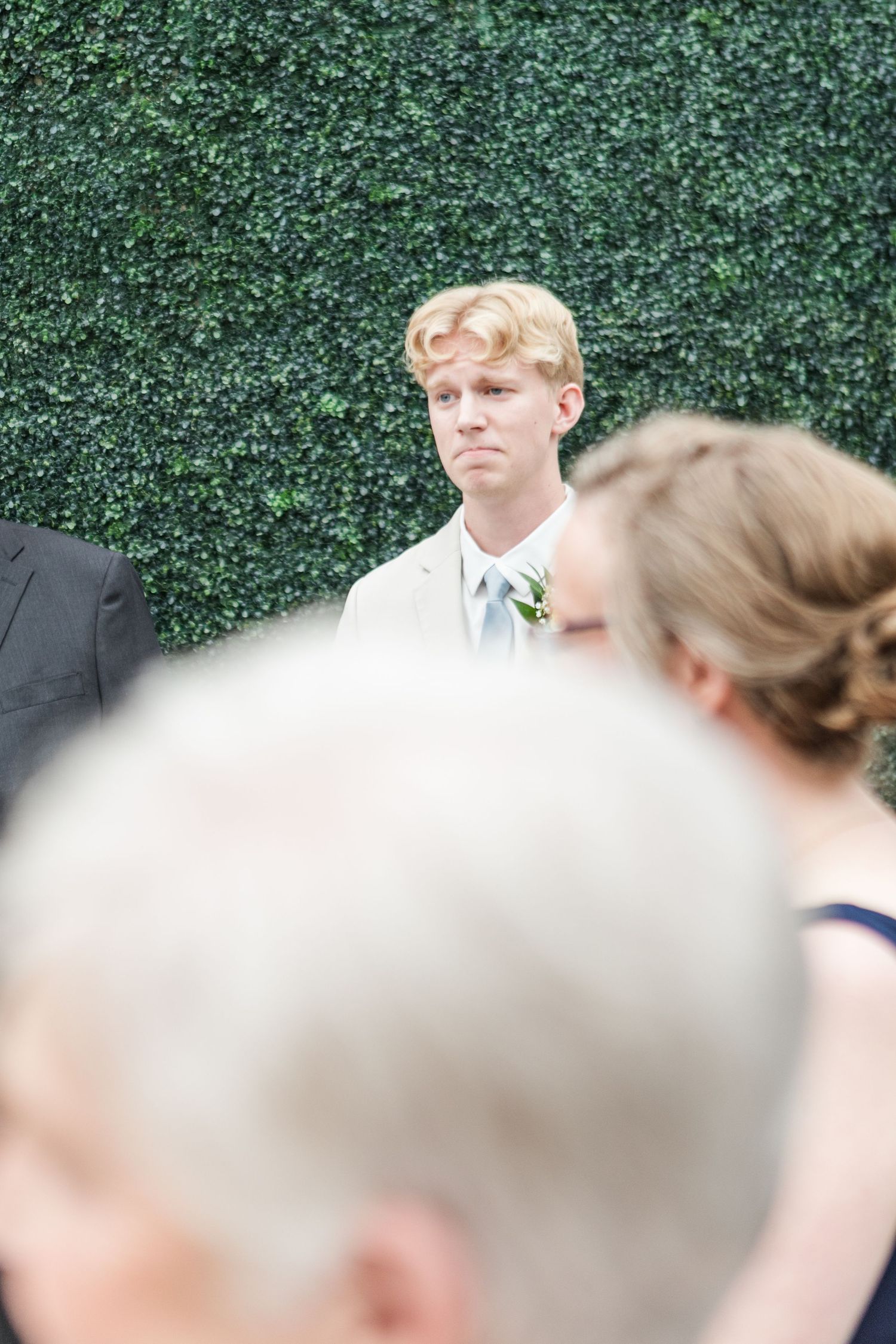 Wedding guests stand in front of a green hedge wall at an outdoor ceremony venue.
