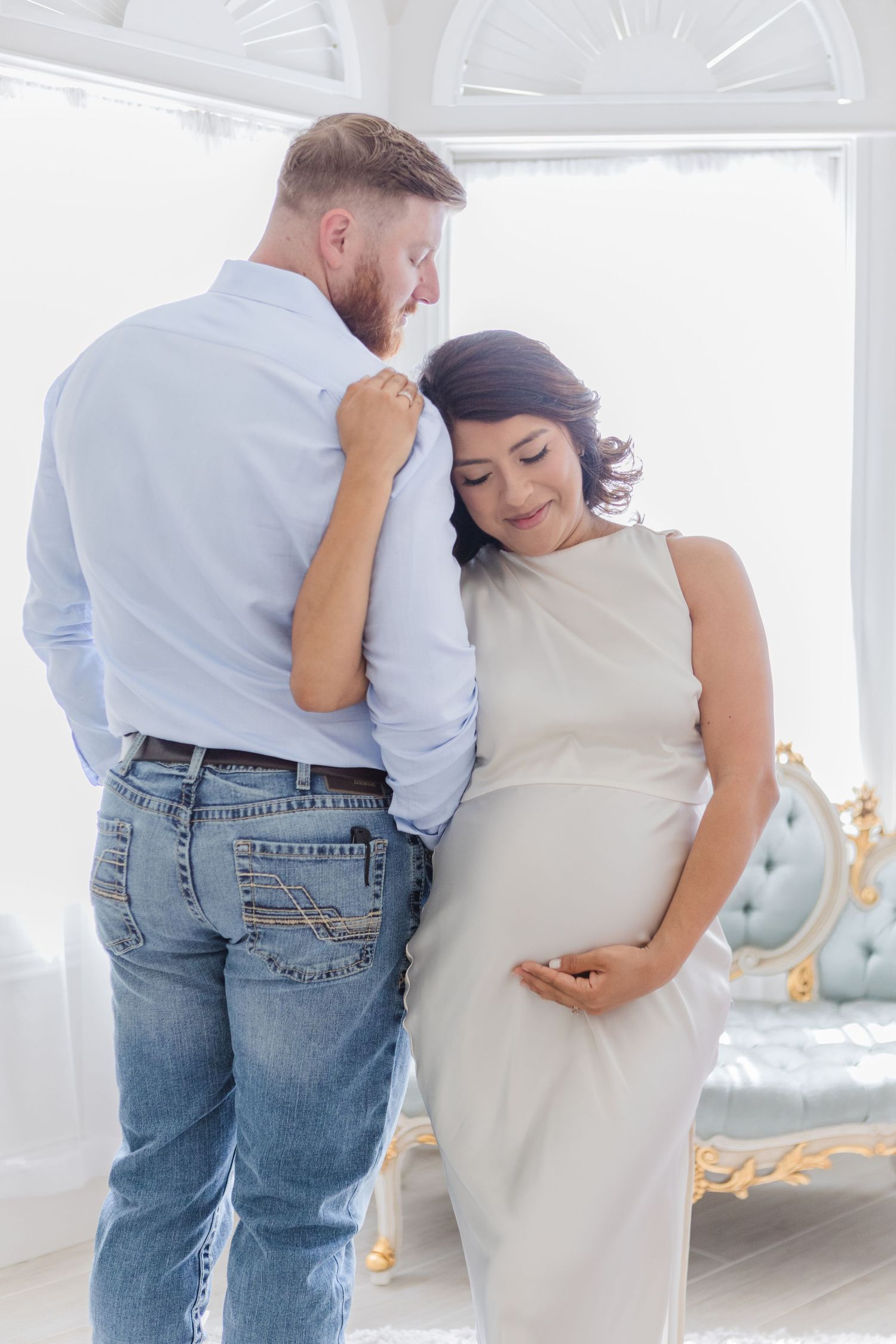 Intimate maternity portraits in white room with couple embracing and touching belly.