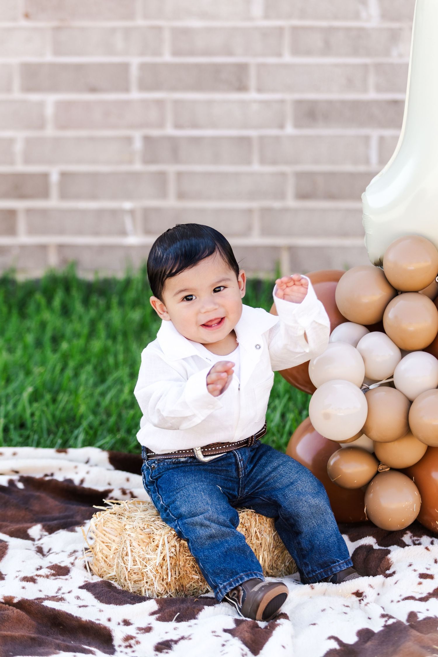 Smiling toddler in casual outfit sits on hay bale with balloon backdrop.