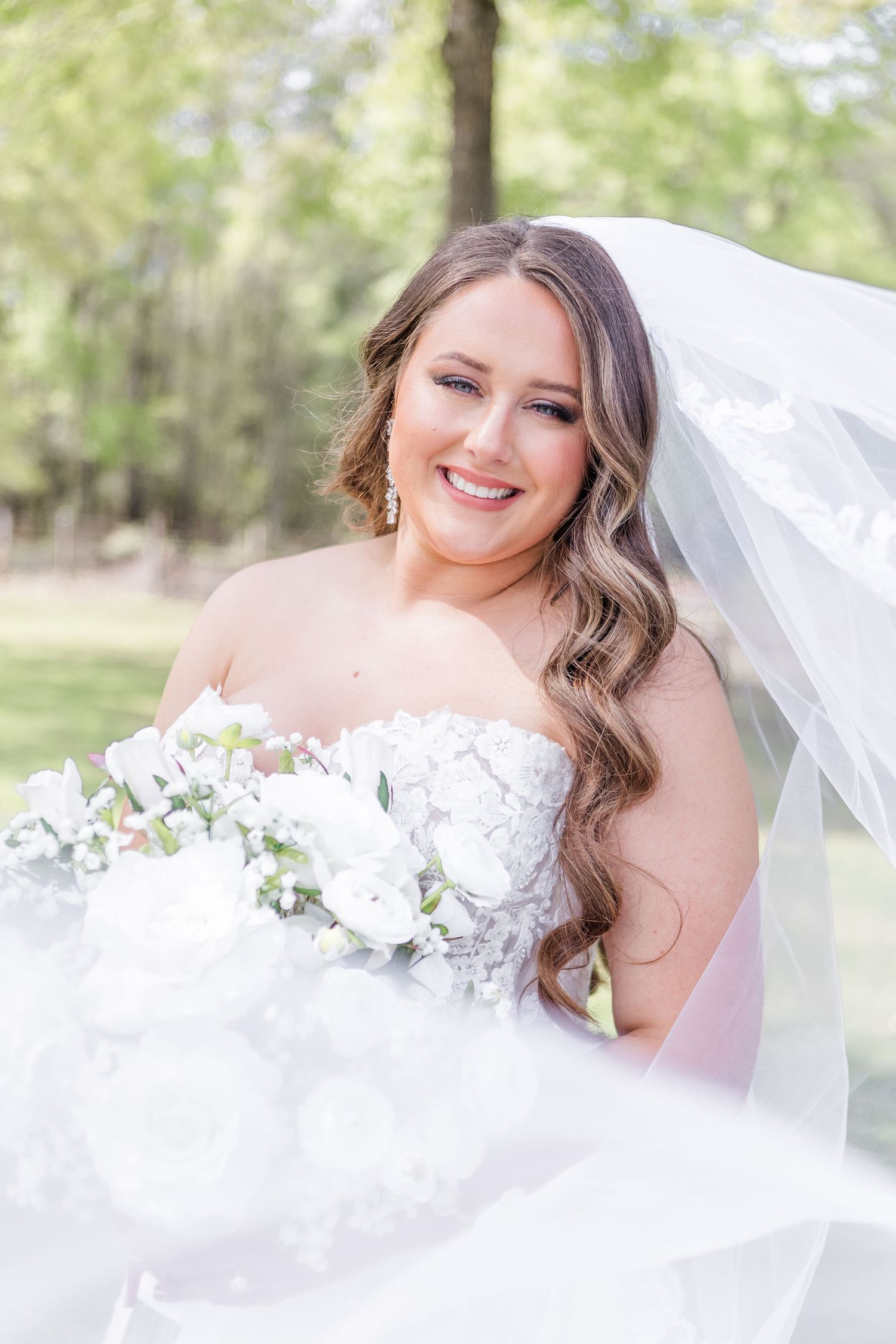Elegant bride holding white bouquet and wearing lace dress with flowing veil outdoors.