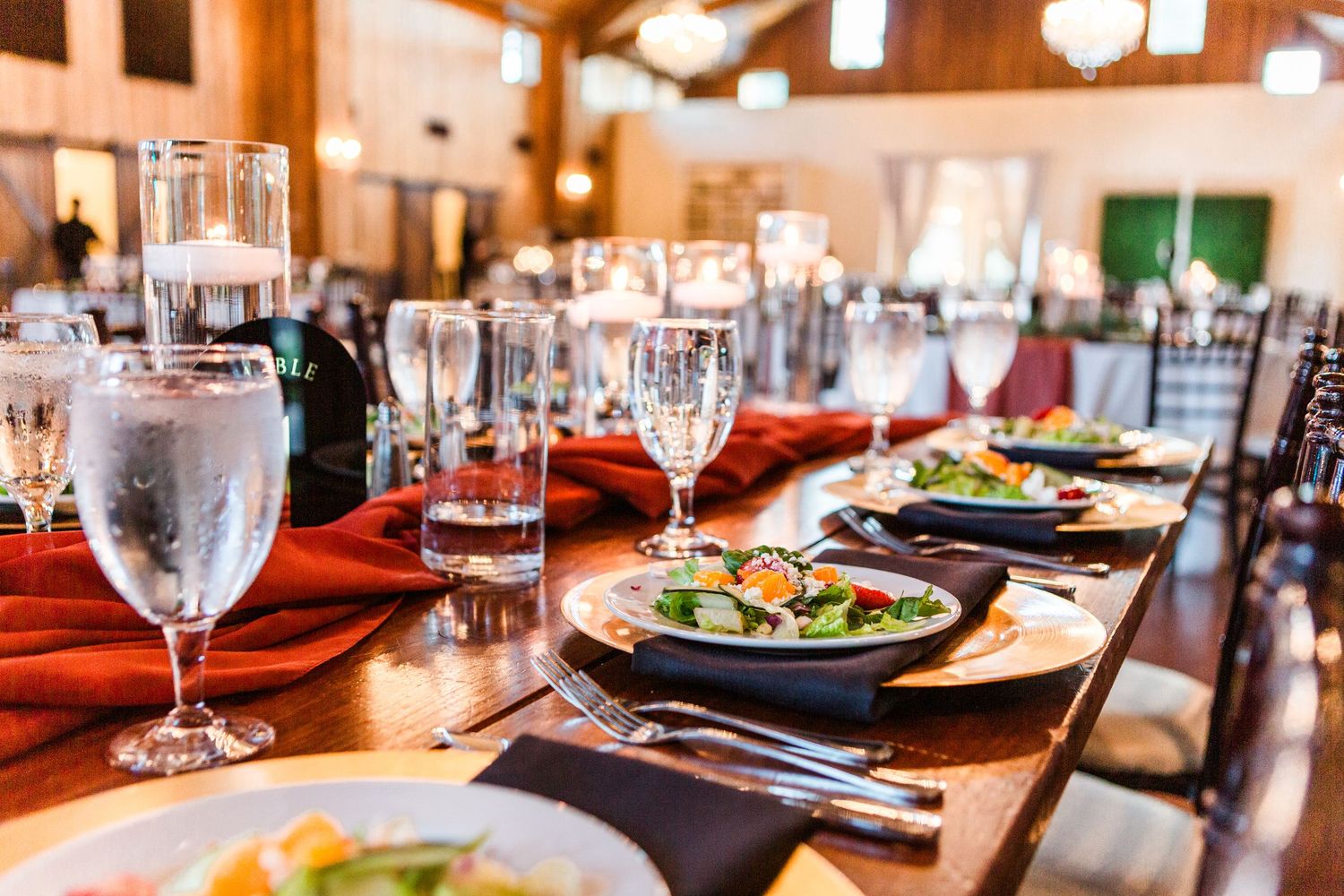 Wedding reception tables set with red accents, glassware and fresh greenery in a rustic venue.