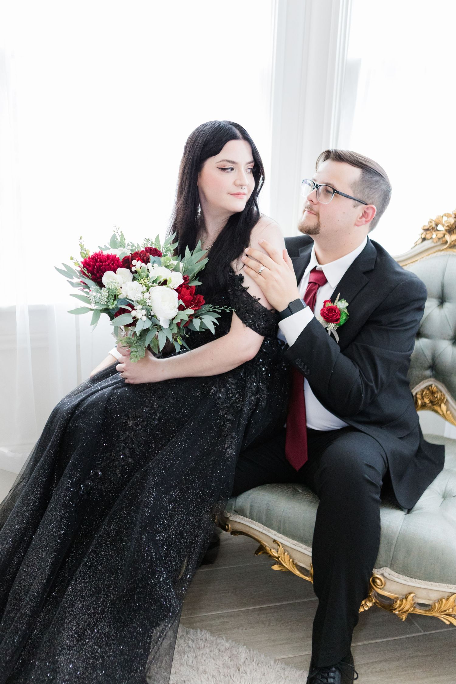 Couple shares romantic moment on vintage settee with red and white floral bouquet.