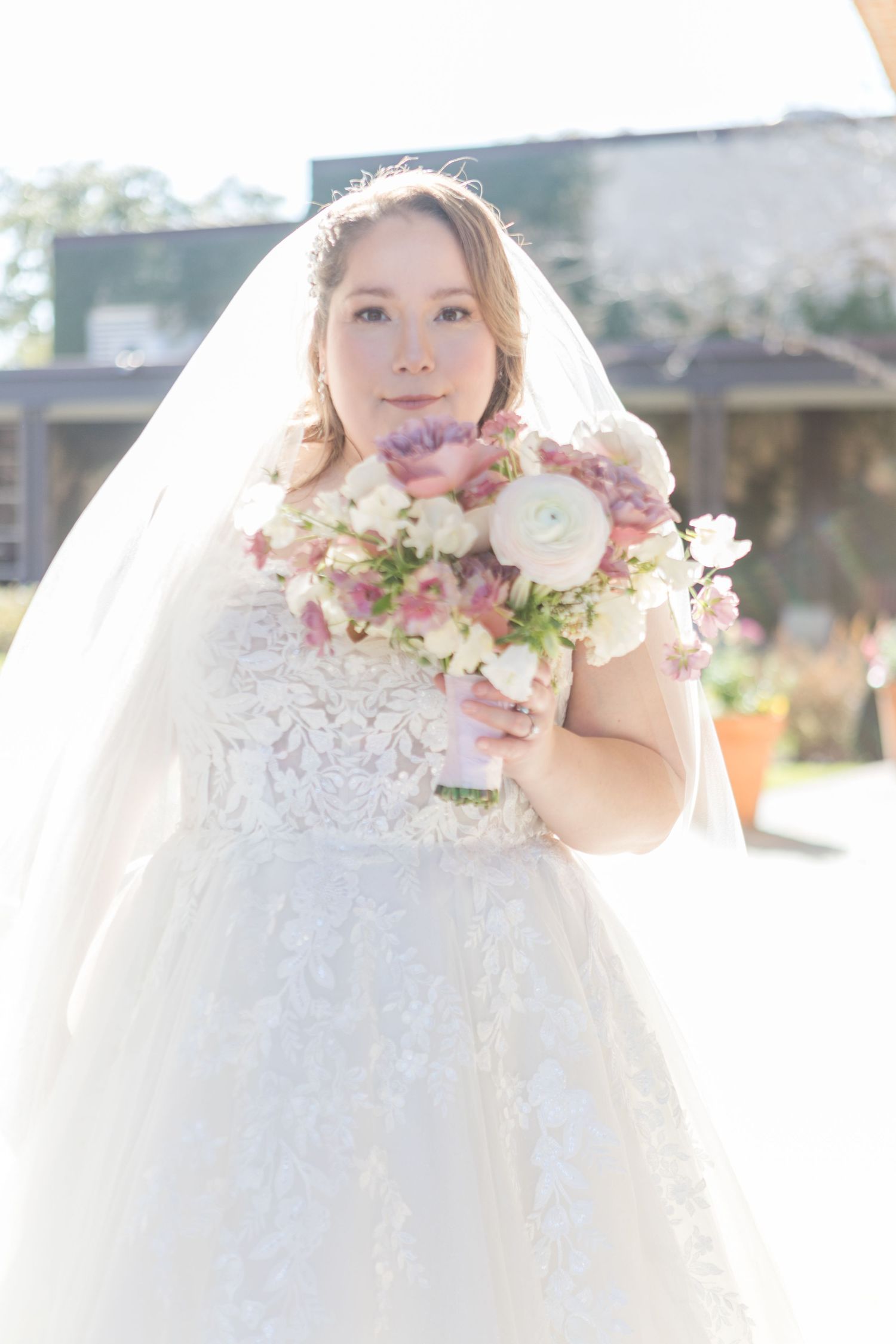 Elegant bride in lace gown with soft pink and white floral bouquet against sunlit backdrop.