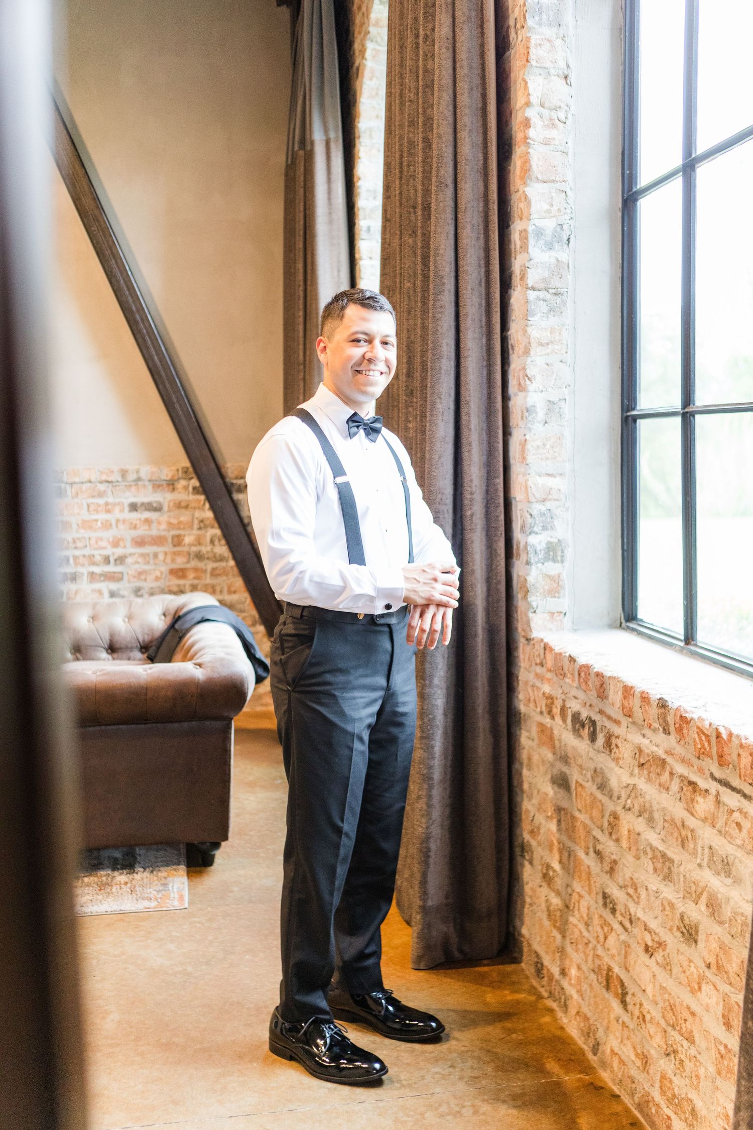 Wedding party member in formal attire standing by exposed brick wall and windows.
